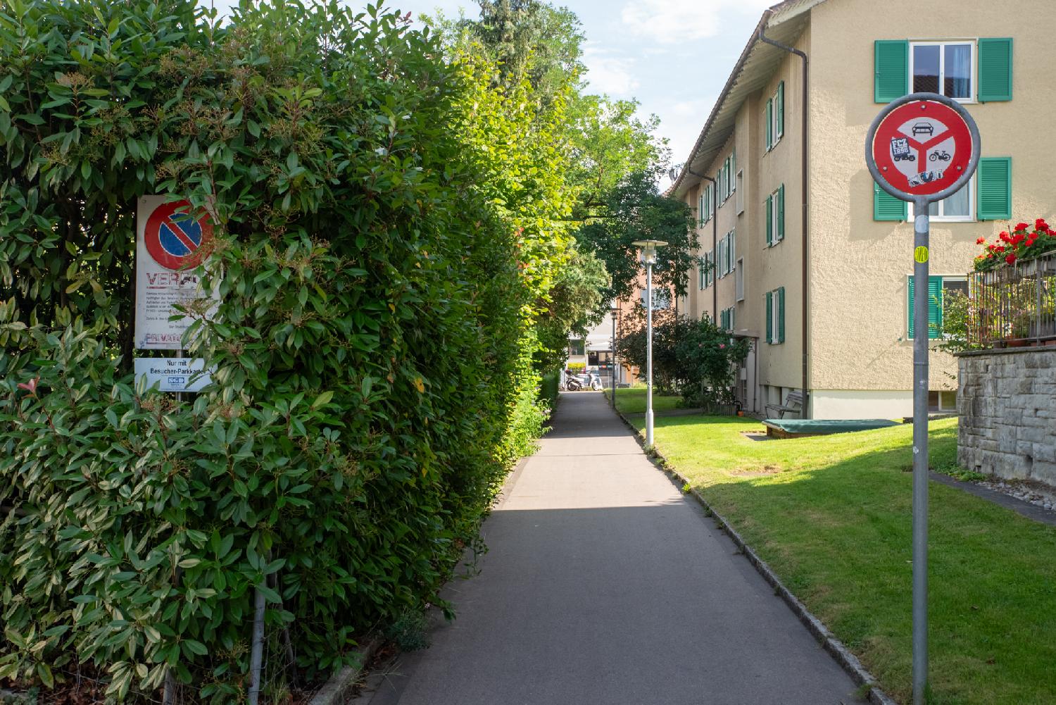 A pedestrian alley with green hedges on the left side and a 3-storey yellow building with green blinds on the right side. Signs in the foreground indicate that it is forbidden to enter or park with any motorized vehicule.