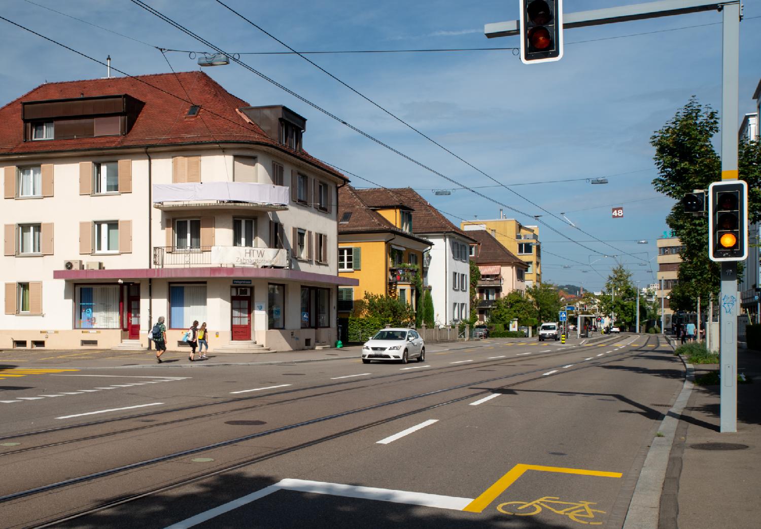 A road with a tram line in the middle and car lines on each side. There are 3-4 storey-buildings on each side of the road; a bike path is marked on the right side of the street. There's an orange traffic light on the right.