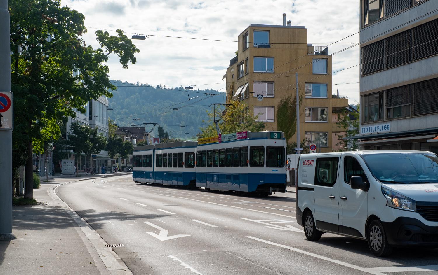 A white and blue tram on a street that has two lines on the left of it and one line on the right. Multiple buildings and a tram stop are visible, as well as the Uetliberg in the background.
