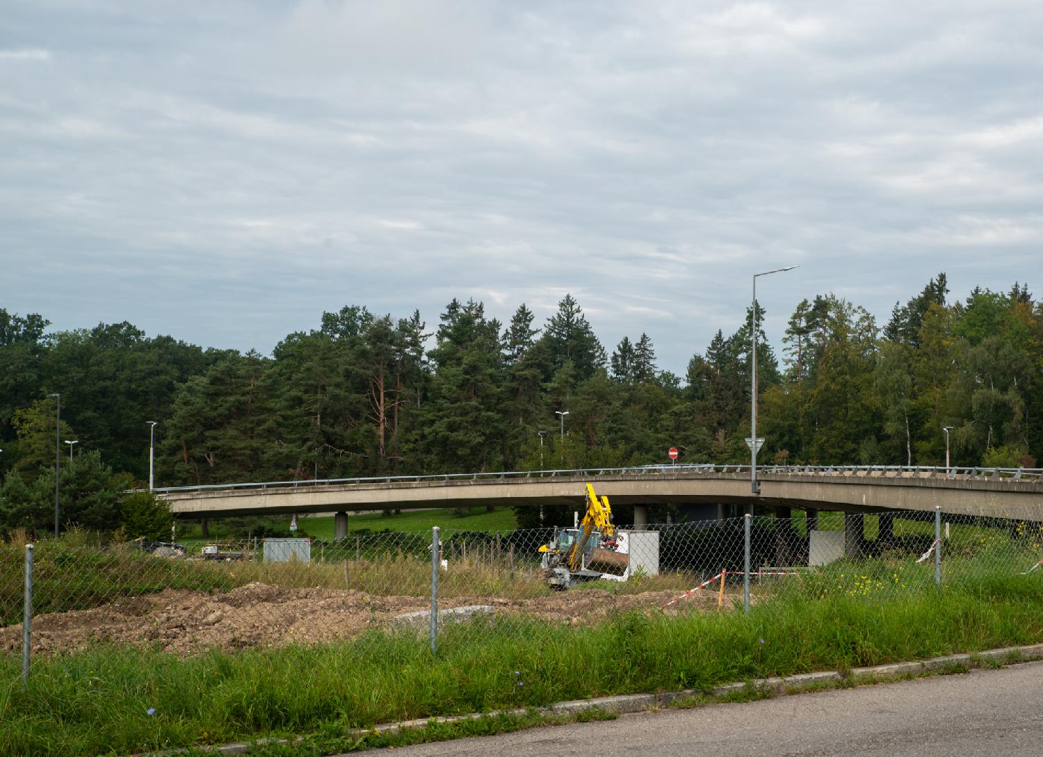 A curved road forming a bridge above a piece of land. There are trees behind the road, and a yellow excavator on the land below the road.