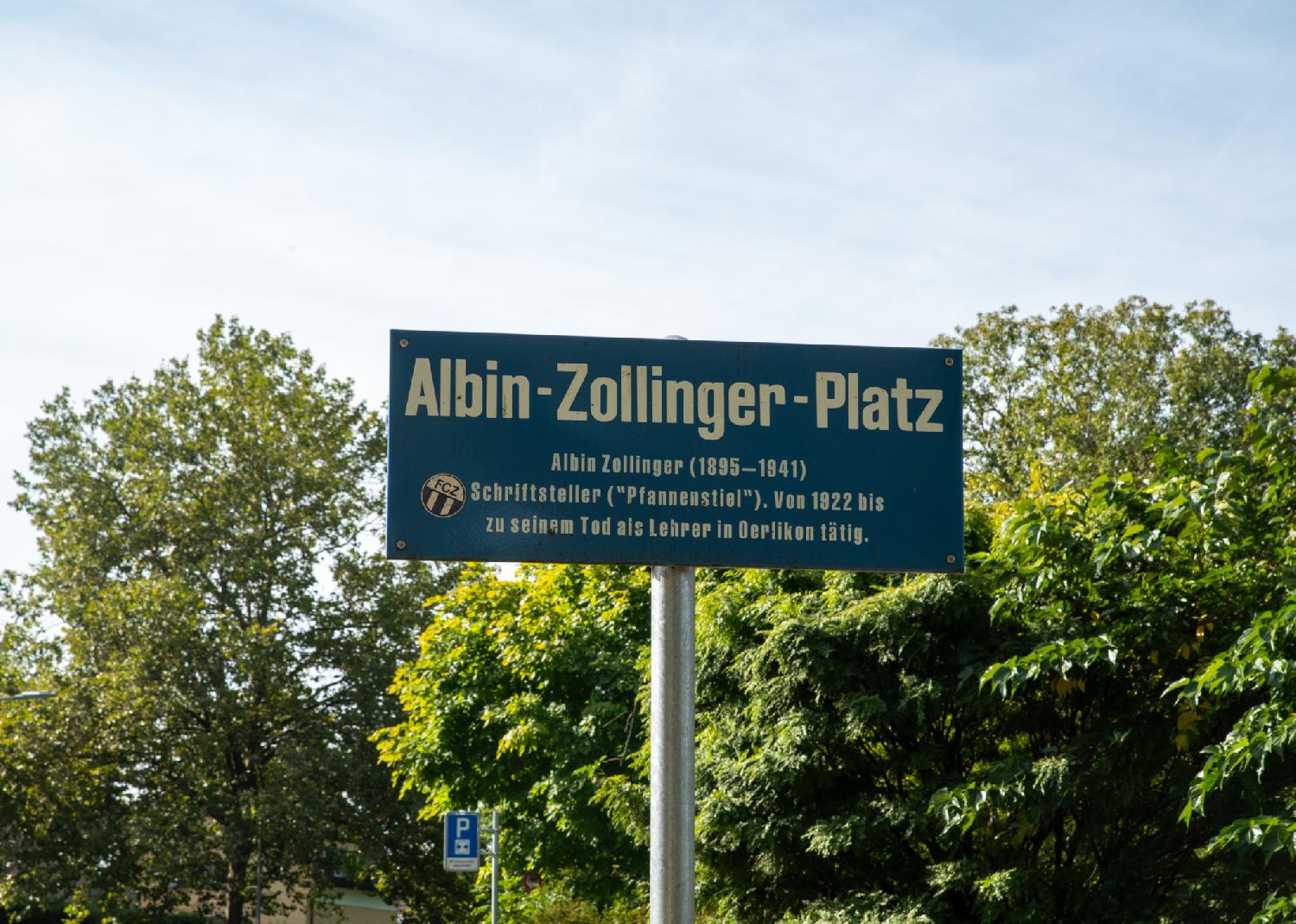 A white on blue street sign attached to a pole, with trees in the background. The sign reads "Albin-Zollinger-Platz (1895-1941) - Schriftsteller ("Pfannenstiel"). Von 1922 bis zu seinem Tod als Lehrer in Oerlikon tätig".