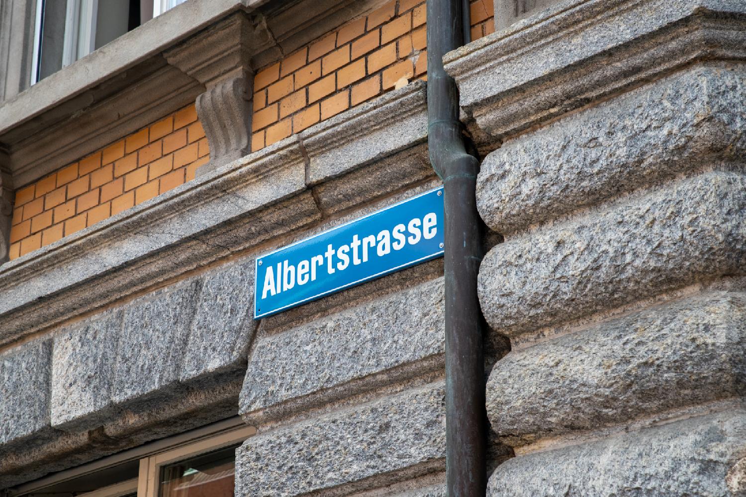 A blue street sign with Albertstrasse written in white, riveted to a stone wall next to a gutter and below four ranks of orange bricks.