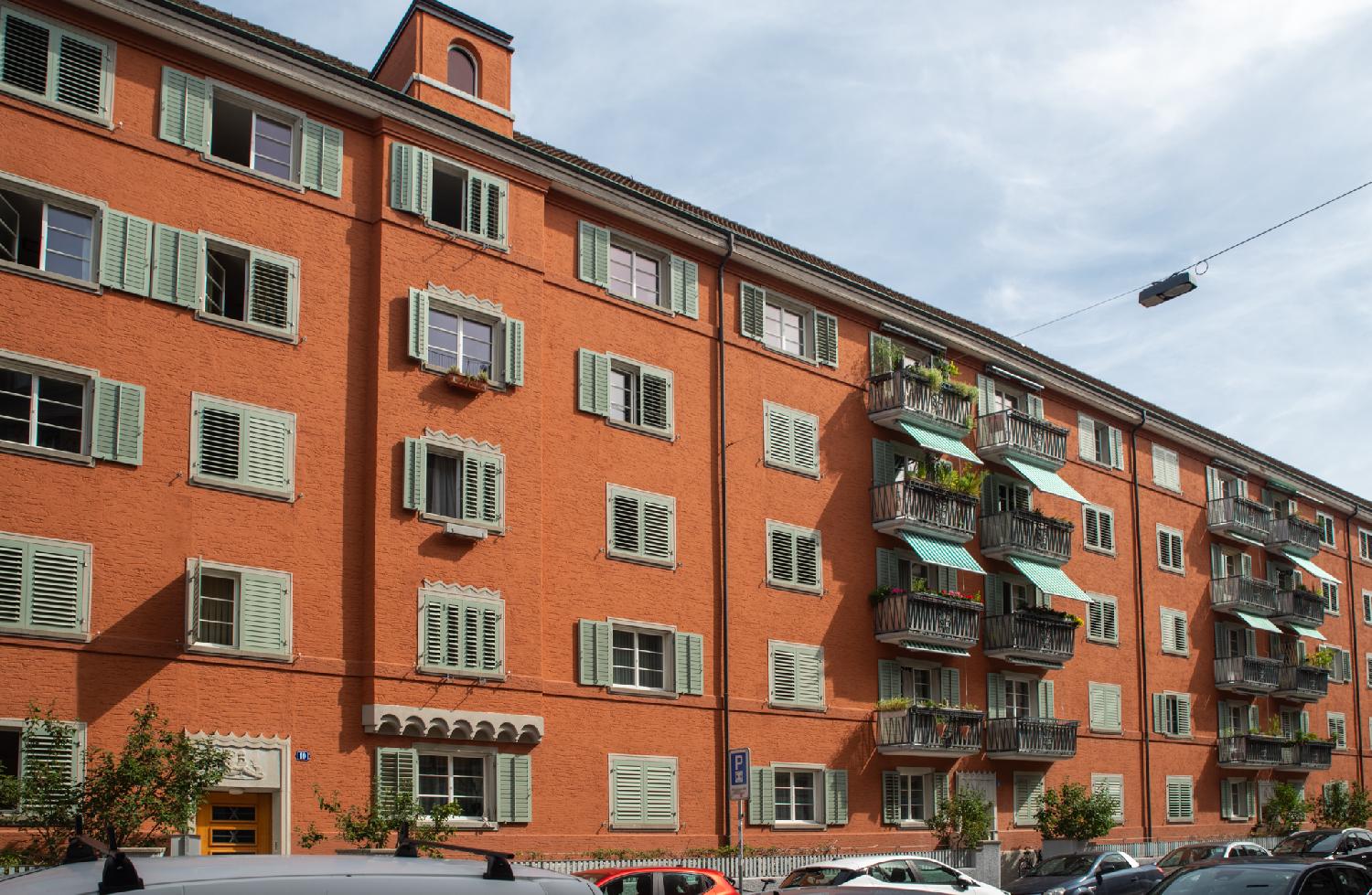 A group of three 5-storeys orange buildings with light green shutters at all windows, some of them open, some of them closed. The two buildings on the right also have balconies with green awnings.