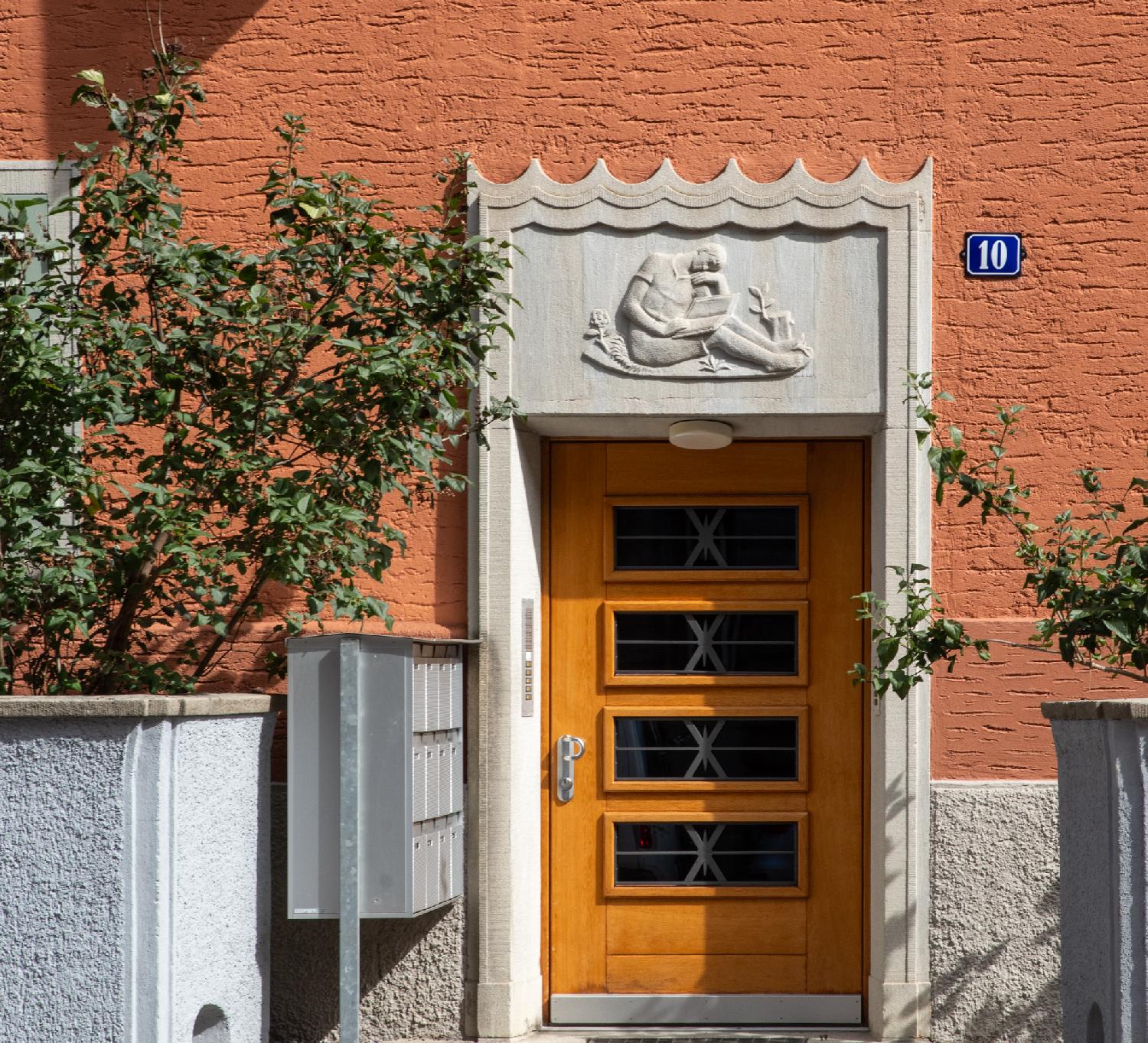 A wooden and glass door with, above it, a stone bas-relief representing a sitting person that looks bored or asleep while reading a book. There are metallic mailboxes on the left of the door.