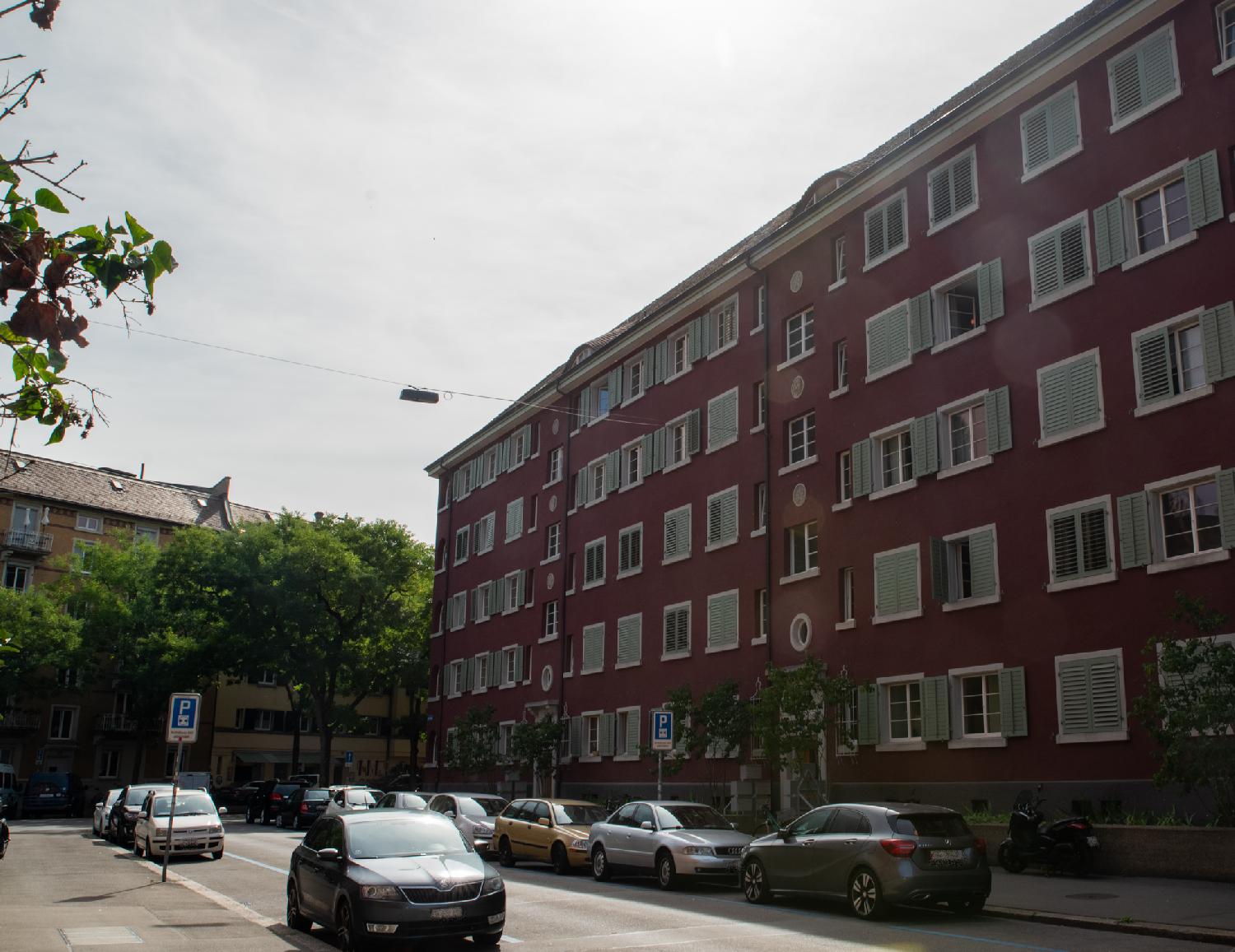 A group of three adjacent 5-storey dark red buildings with pale green blinds, on a street with cars parked on each side.