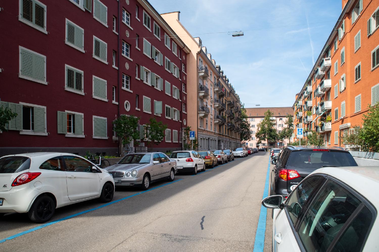 A street with cars parked on blue parking spots on both sides of the street. There are 5-storey buildings of different shades of orange/red/peach on both sides of the street and at the street intersection in the background.