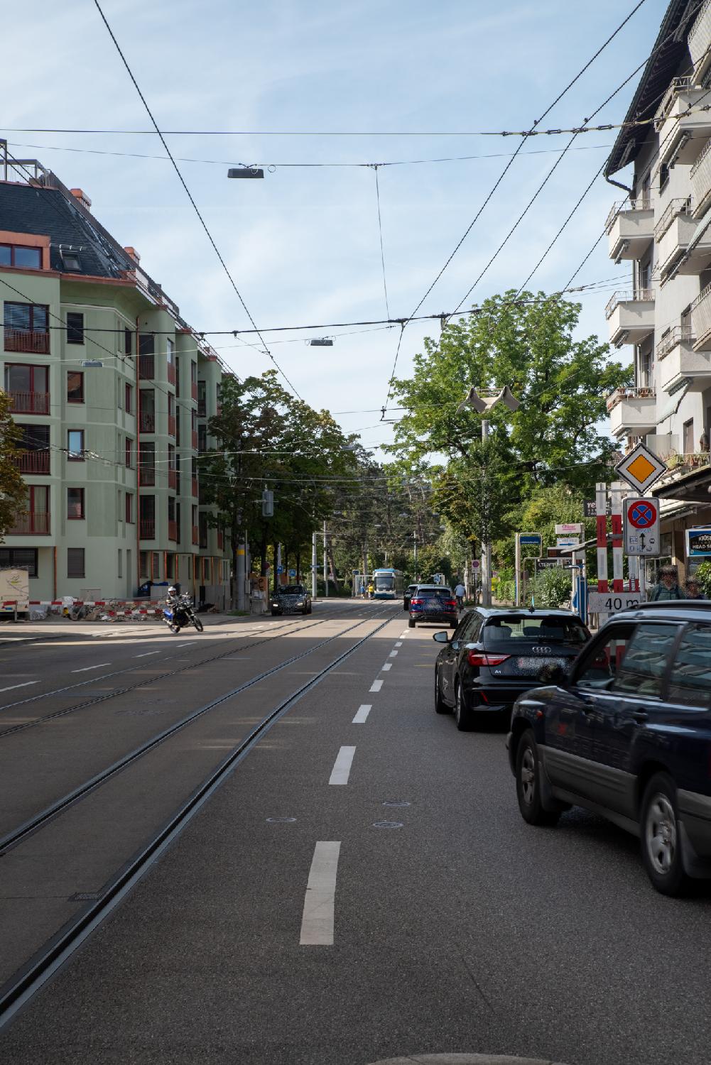 A road with a tram line in the middle and car lines on each side. There are trees on the sidewalks and 3-4-storey buildings behind the trees. A bus stop and a tram stop are visible on the picture, with a tram stopped at the tram stop.