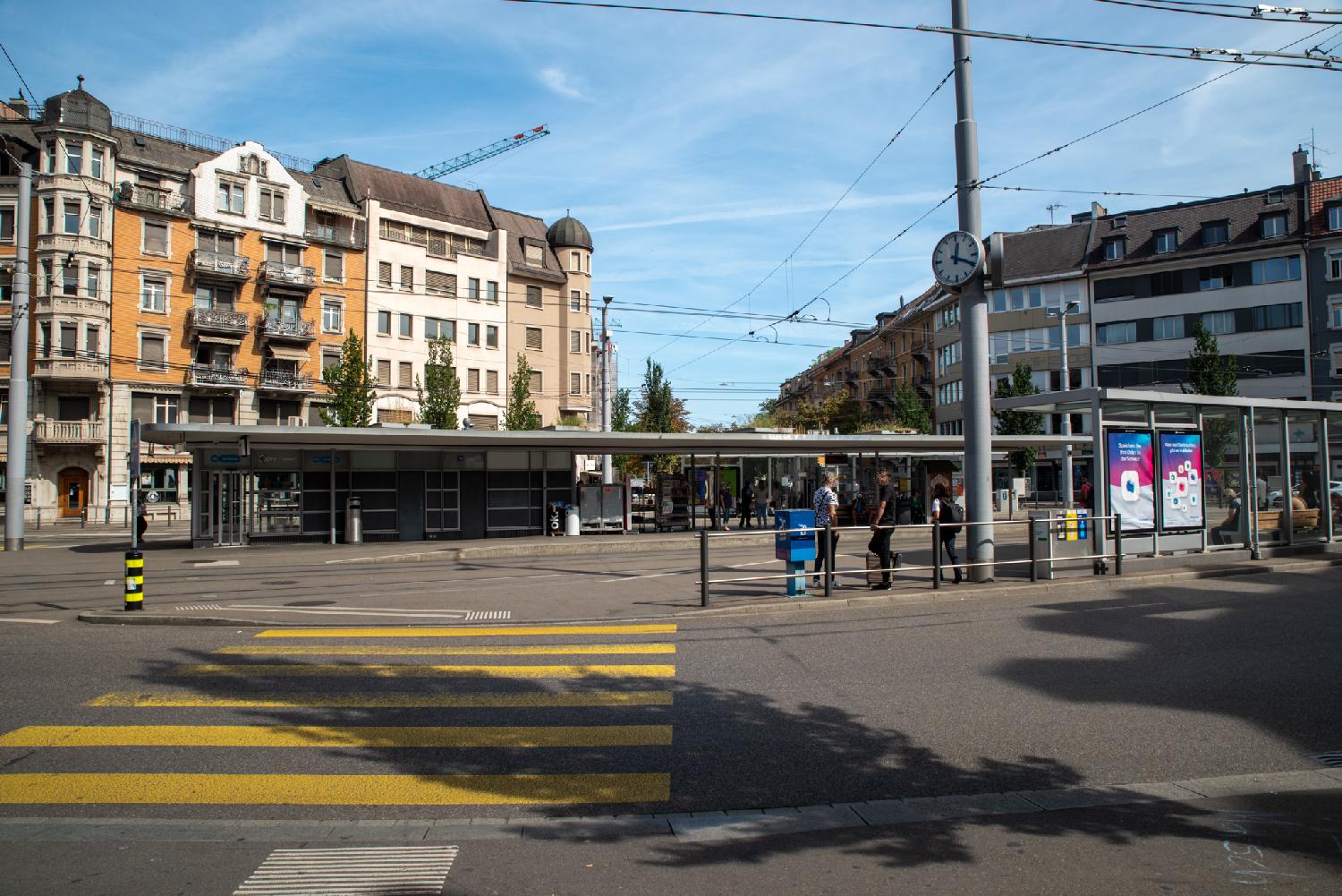 A large tram stop in the middle of a town square. Behind the tram stop, two blocks of buildings, one with bay windows and balconies, another one more plain, and a street going away from the square. On the foreground, a yellow zebra crossing leading to the tram stop.