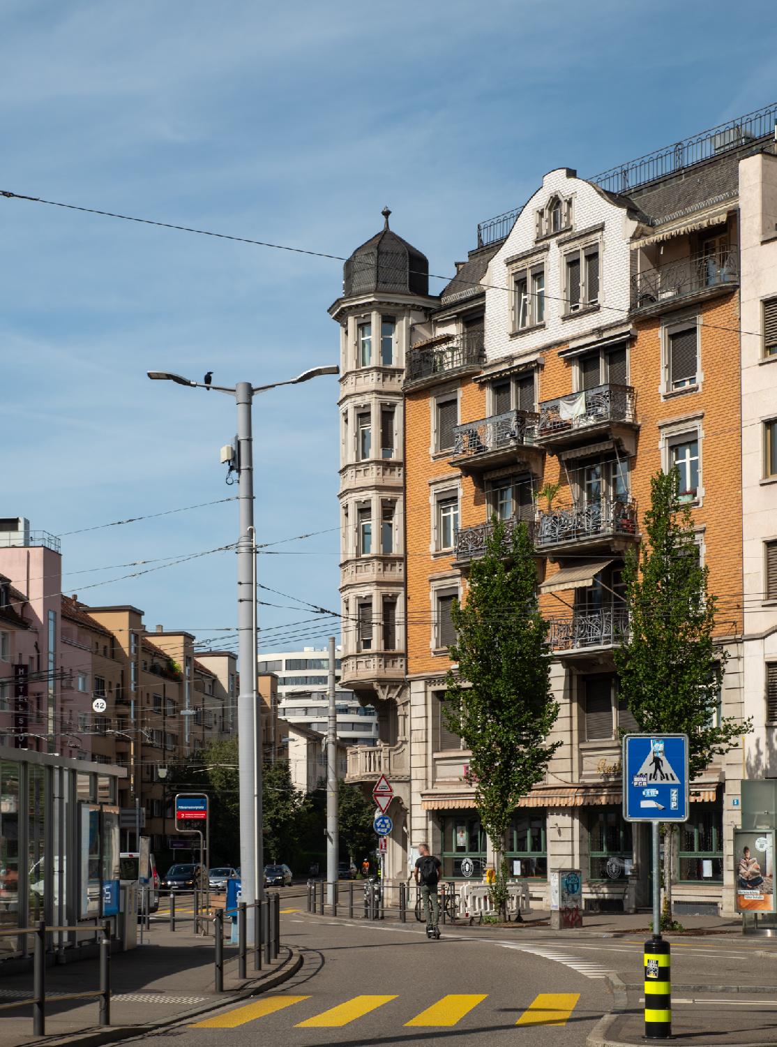 Curve on a town square with, on the left, a tram stop with the tram 2 sign visible; on the right a salmon building with octogonal bay windows on the corner of the street. In the foreground, a yellow zebra crossing crossing the street.
