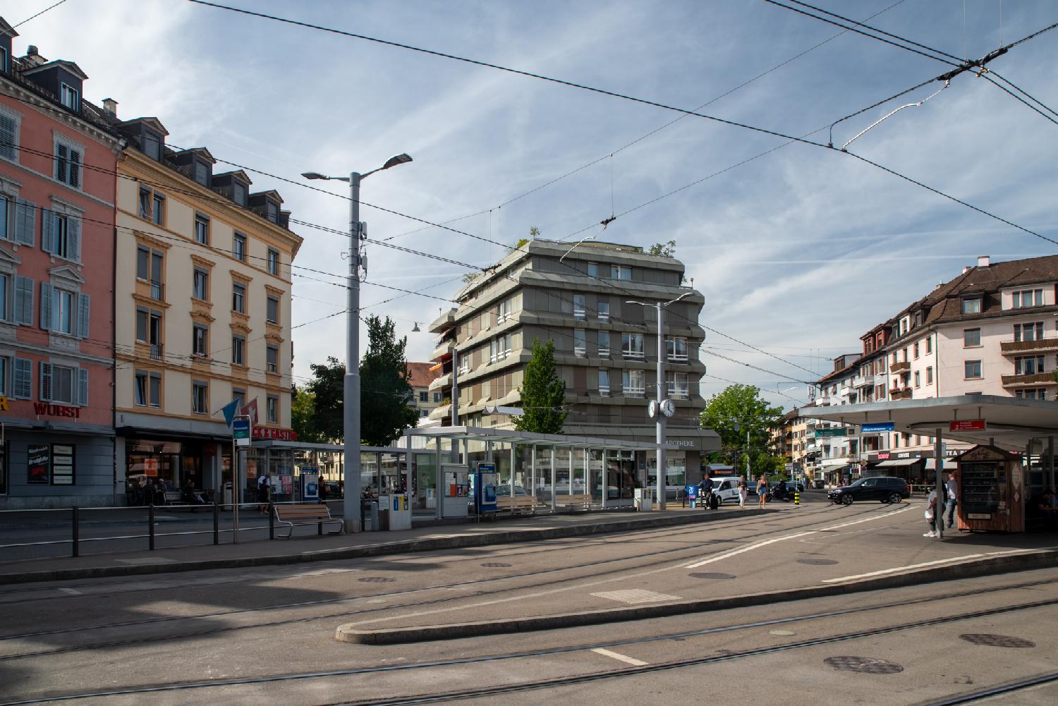 A tram stop at a traffic intersection/square with multiple rail tracks. The background shows two streets arriving on the square. The tram stop shows signs for lines 2 and 3; the one on the left side has three wooden benches and a ticket distributor. The one on the right has a food distributor in the shape of a chalet.
