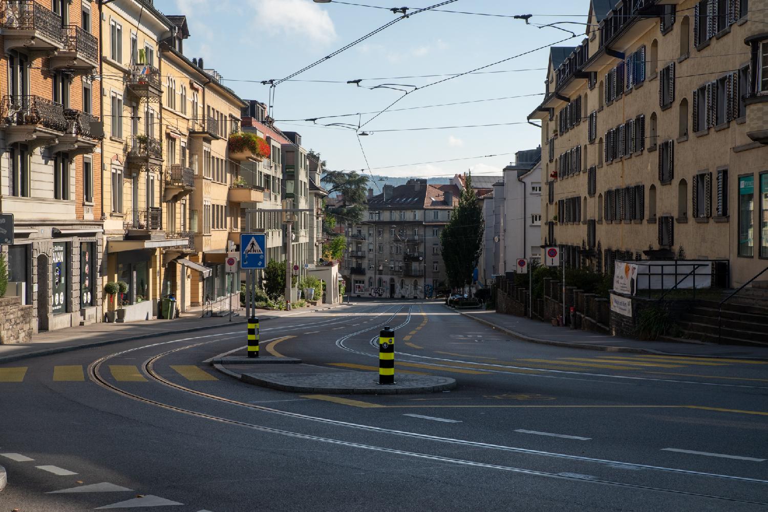 A curve in a 2-way street with 3-4-storey buildings on each side, and two tram tracks in the middle. The street is going down.