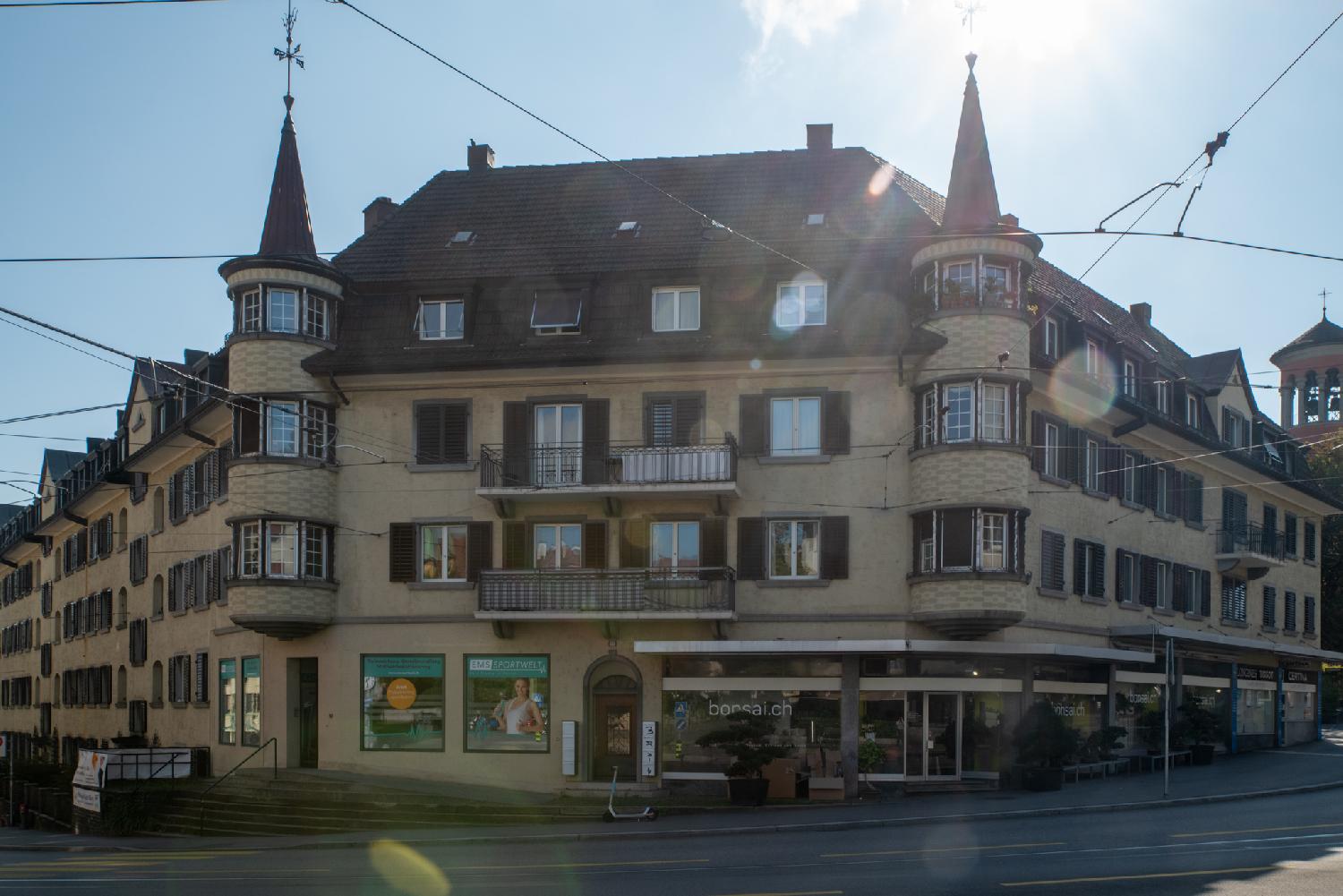 A four-storey building at a street angle with shops on the ground floor (a bonsai florist and a fitness studio) and two small tower-like structures on the side, with bay windows and spires.