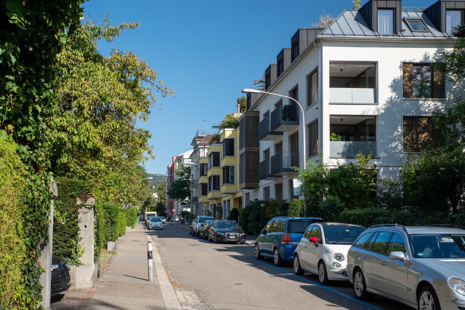 A residential street that shows 3-4 story-buildings on its right and green hedges and trees on its left. A lot of cars are parked on blue parking spots on the street.