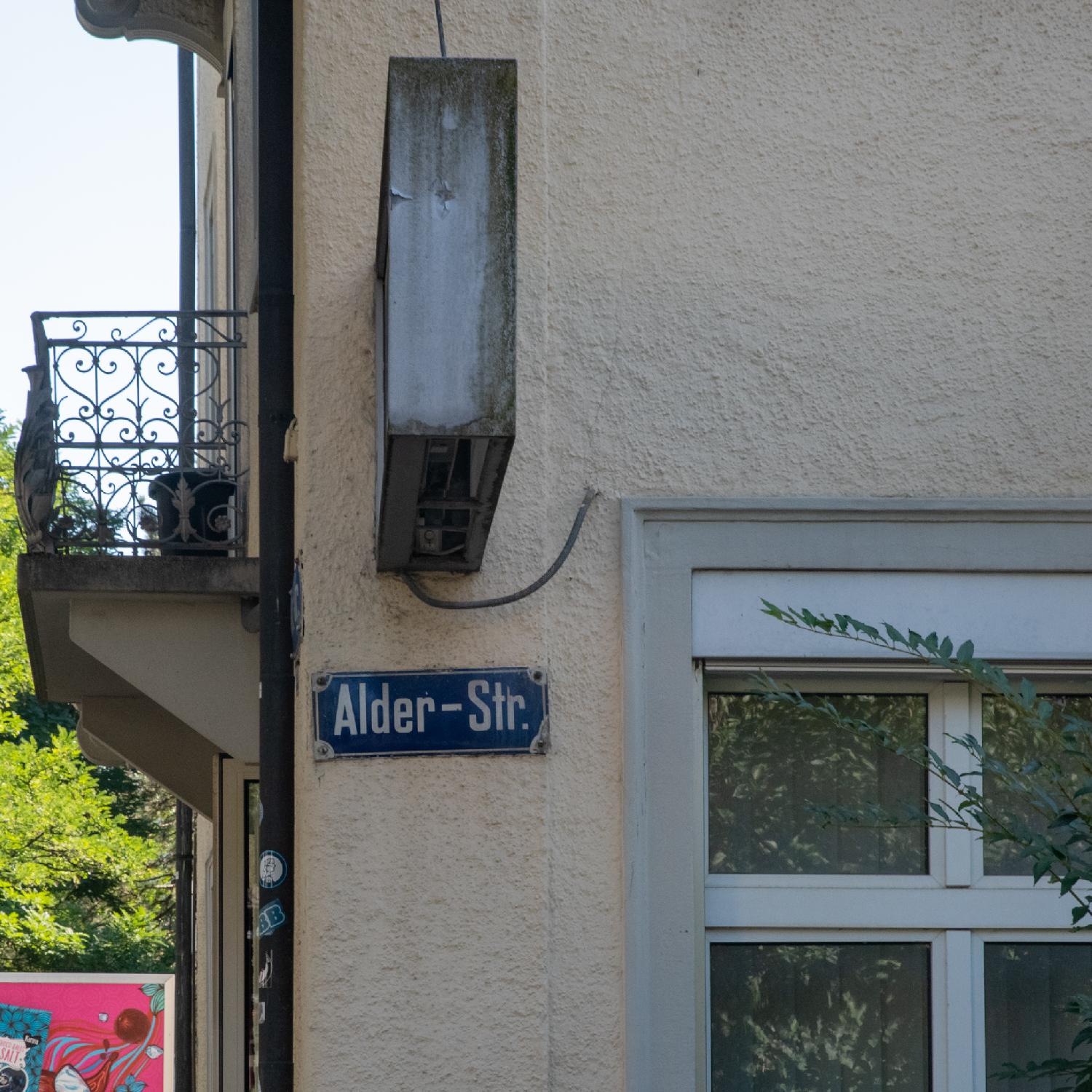 A blue street sign, written "Alder-Str.", on a yellow/beige wall with a window and an light panel side. There's a metal balcony on the perpendicular wall.