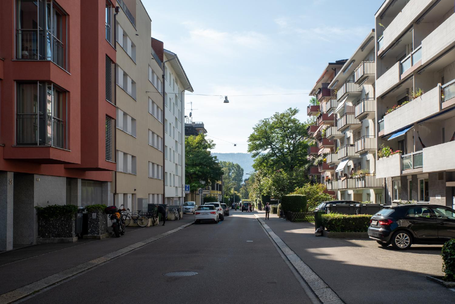 A street with 4-5-story residential or office buildings on each side. There's a visible green hill in the far distance, and some trees/greenery between the end of the street and the hill.