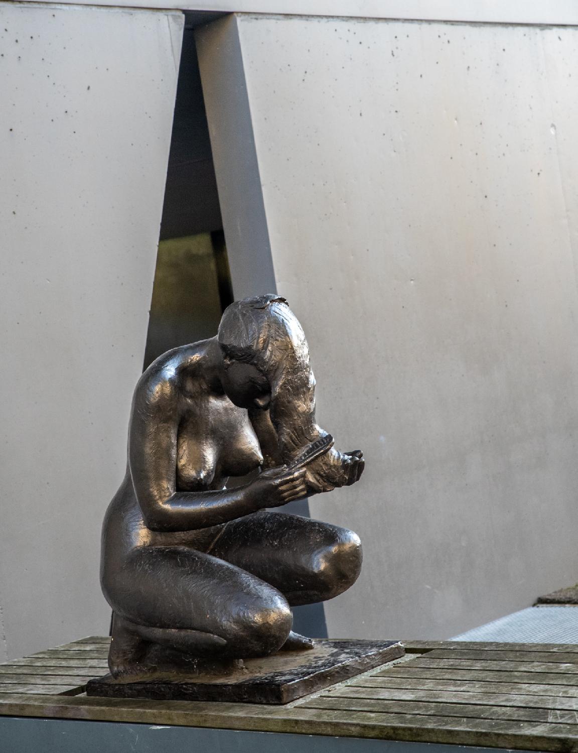 A statue of a naked woman, kneeling and untangling her hair with a comb.