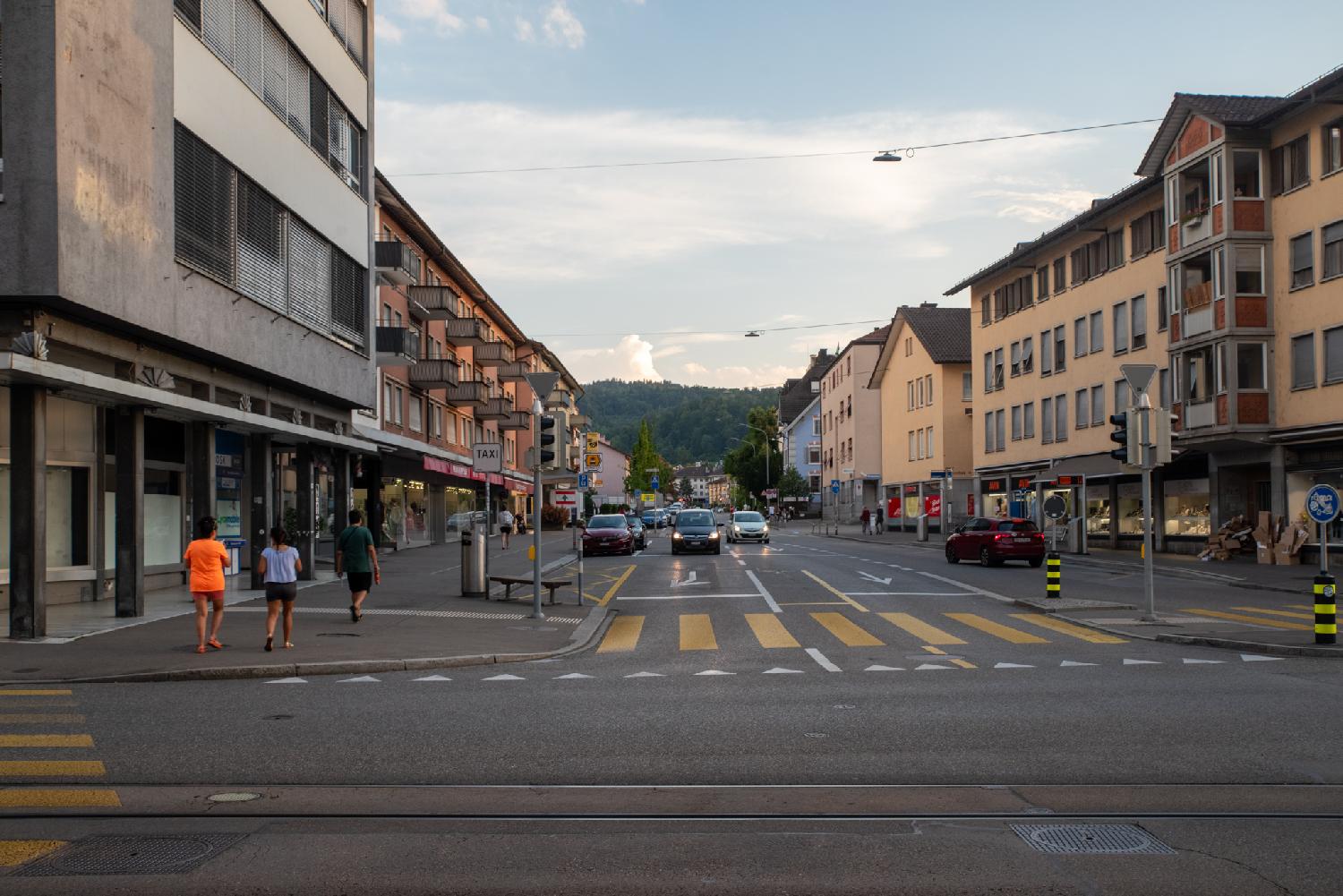 A three-lane, two-way street with 3-4-storey residential buildings with storefronts on the ground floors, a few cars on the street, and the Uetliberg forest hill in the background.