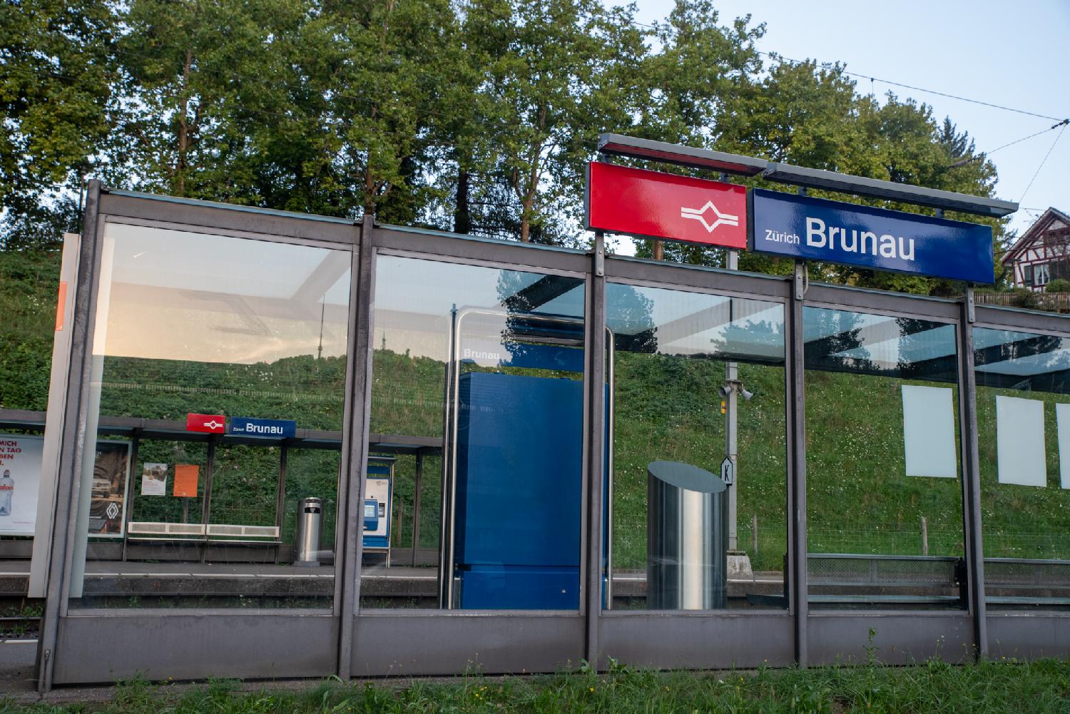Zürich Brunau train station: Two train stops on both sides of train tracks, with a glass shelter (reflecting a green hill), benches and a blue ticket machine.
