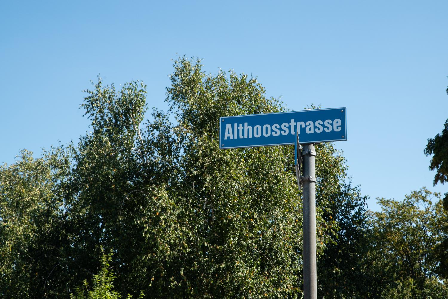 Zürich Althoosstrasse street sign: a blue street sign on a metallic pole in front of low trees and a blue sky