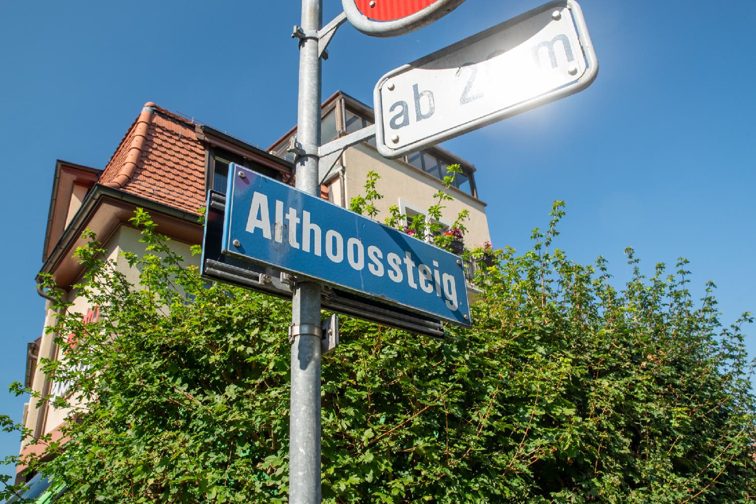 Zürich Althoossteig street name sign: a blue street name sign on a metallic pole, in front of a small tree, in front of a higher residential building wtih a tile roof