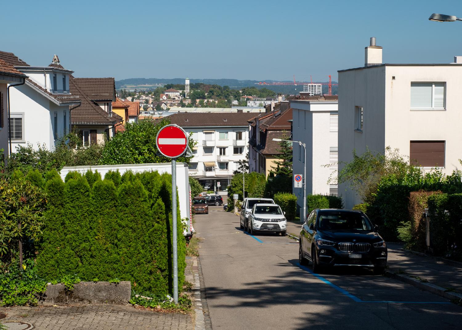 Althoossteig in Zürich: a narrow street, going down, with 3-storey buildings on each side and cars parked on blue parking spots. A red "no entry" sign indicates that the street is going one-way.