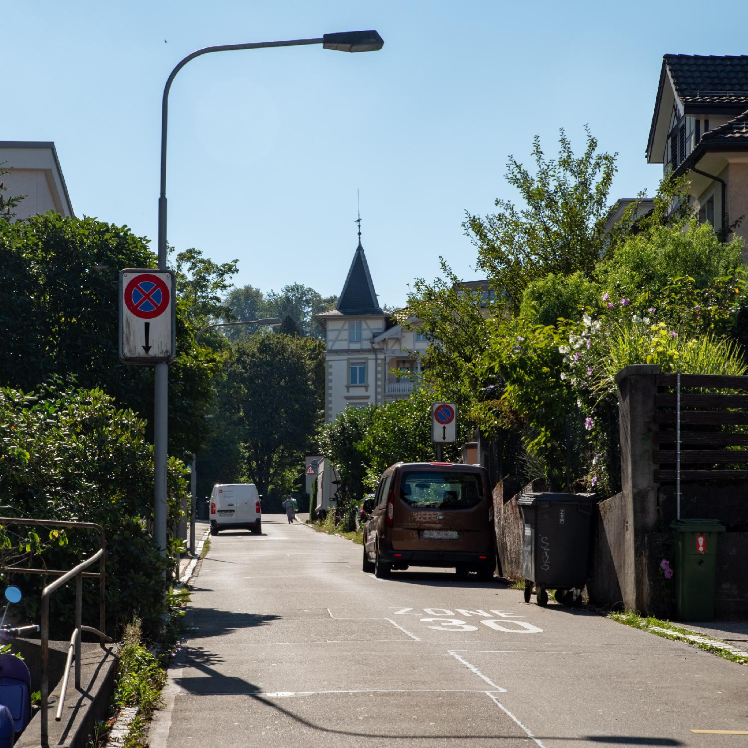 Althoossteig in Zürich: a narrow residential street with a lot of greenery visible, a "Zone 30" sign painted on the street, an a couple of cars parked. In the background of the picture, there is a more imposing building with a spired tower and timber framing, making the corner of the street.
