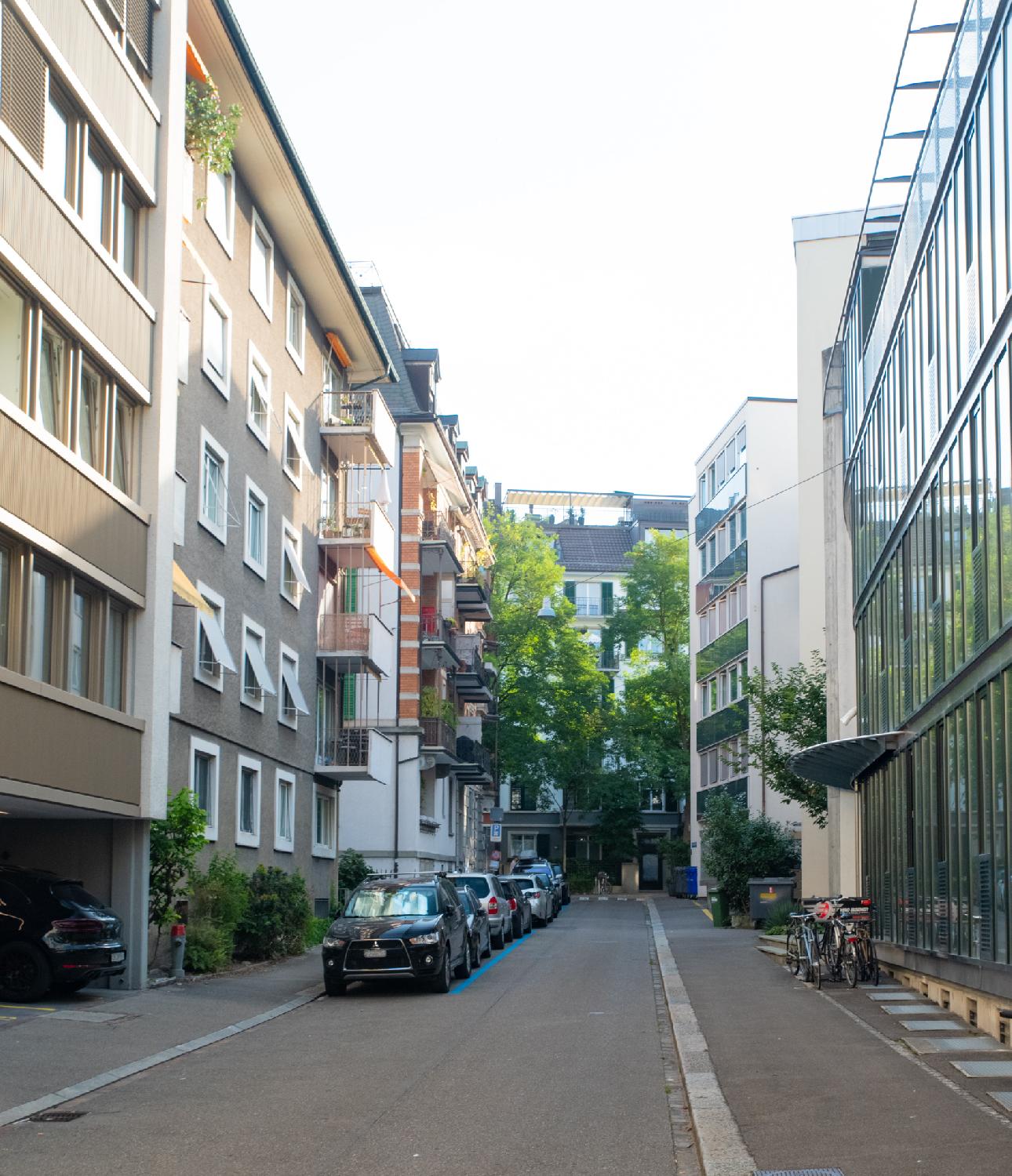 Alte Feldeggstrasse in Zürich: a narrow street with 4-5-story buildings (stone on the left, glass on the right) and cars parked in blue spots on the left
