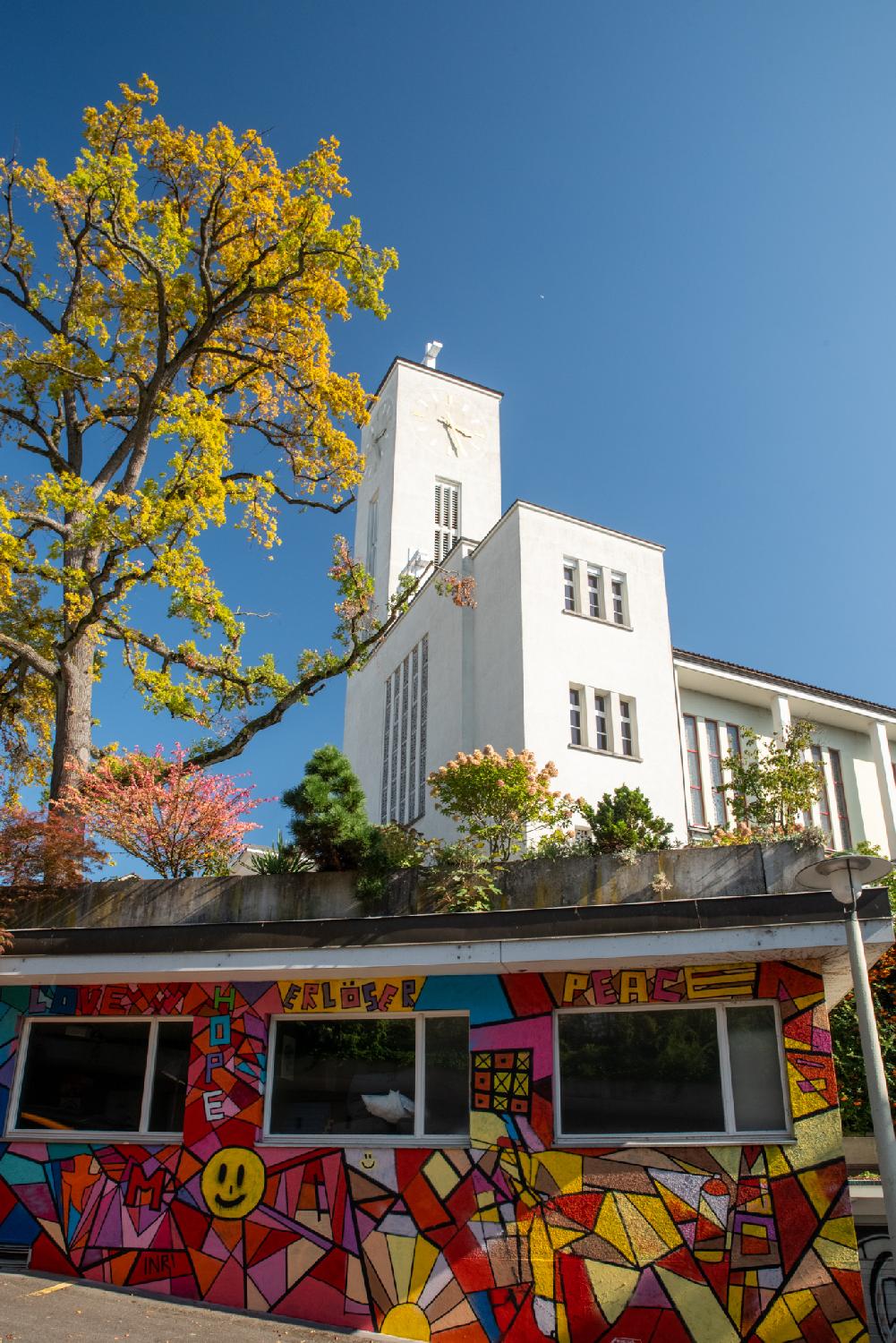 Erlöser Kirche and Jungenhaus in Zürich: a brightly colored low building, with a tree and a white church in the background
