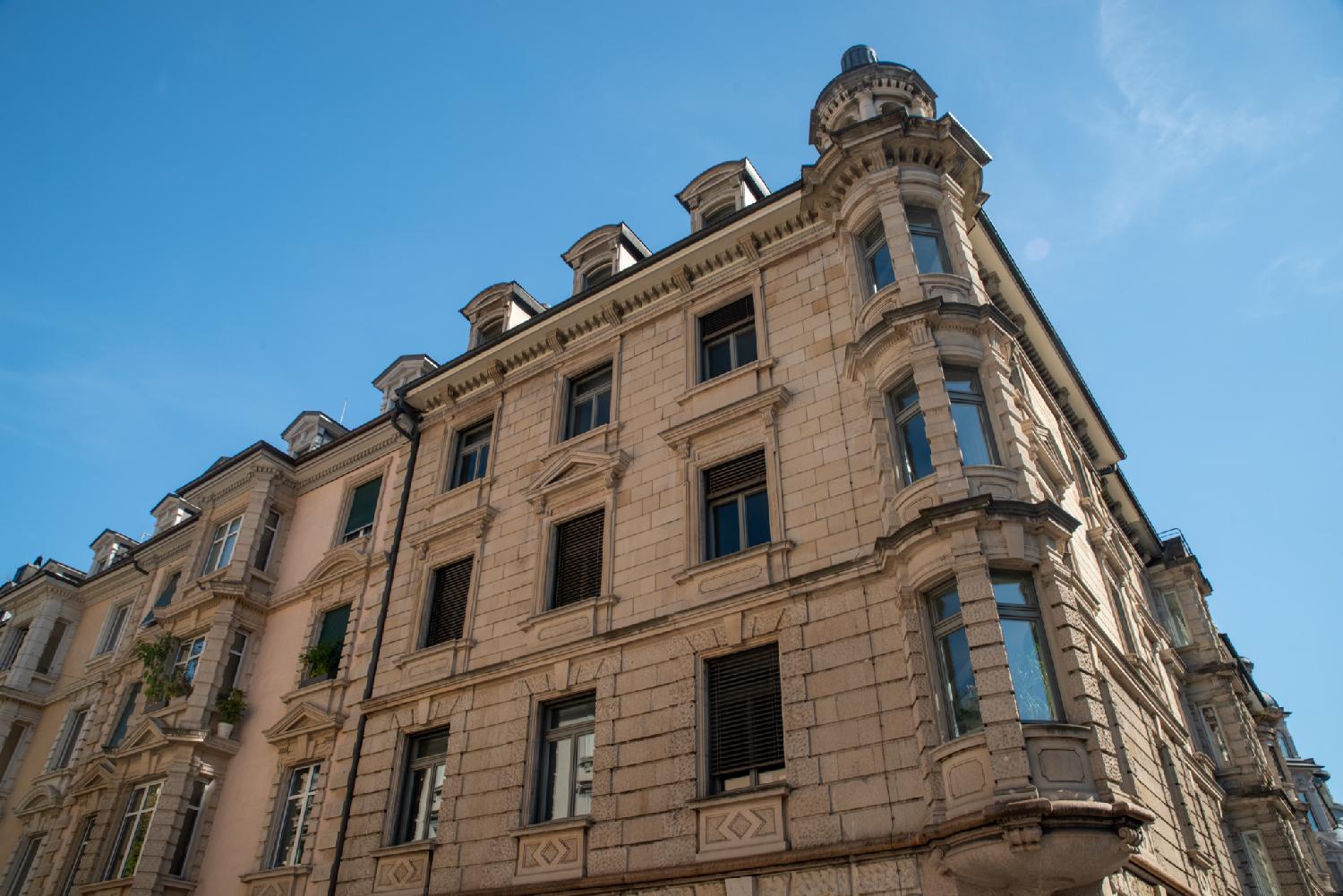 Building at the angle of Alfred-Escher-Strasse and Bodmerstrasse in Zürich. 5-story stone building with angled bay window and roof dormer windows.