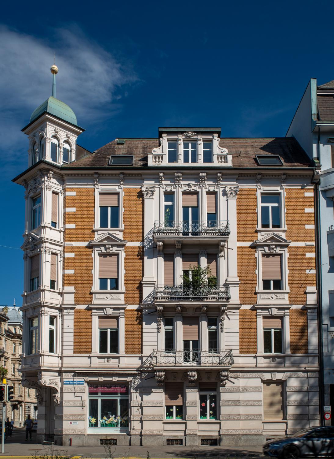 A brown and white 5-story building with square bay window on the street corner, topped by a green roof and a narrow spire. There are storefronts on the ground levels and balconies on the upper levels. The roof floor has a dormer window and a couple of roof windows.