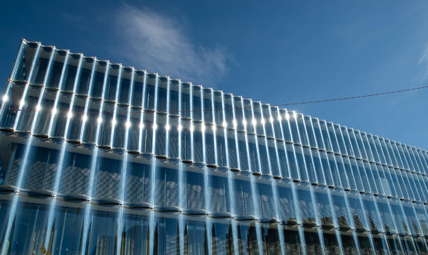 A building made of glass waves, on which the sun reflects multiple times over an arc in the top left corner of the building.