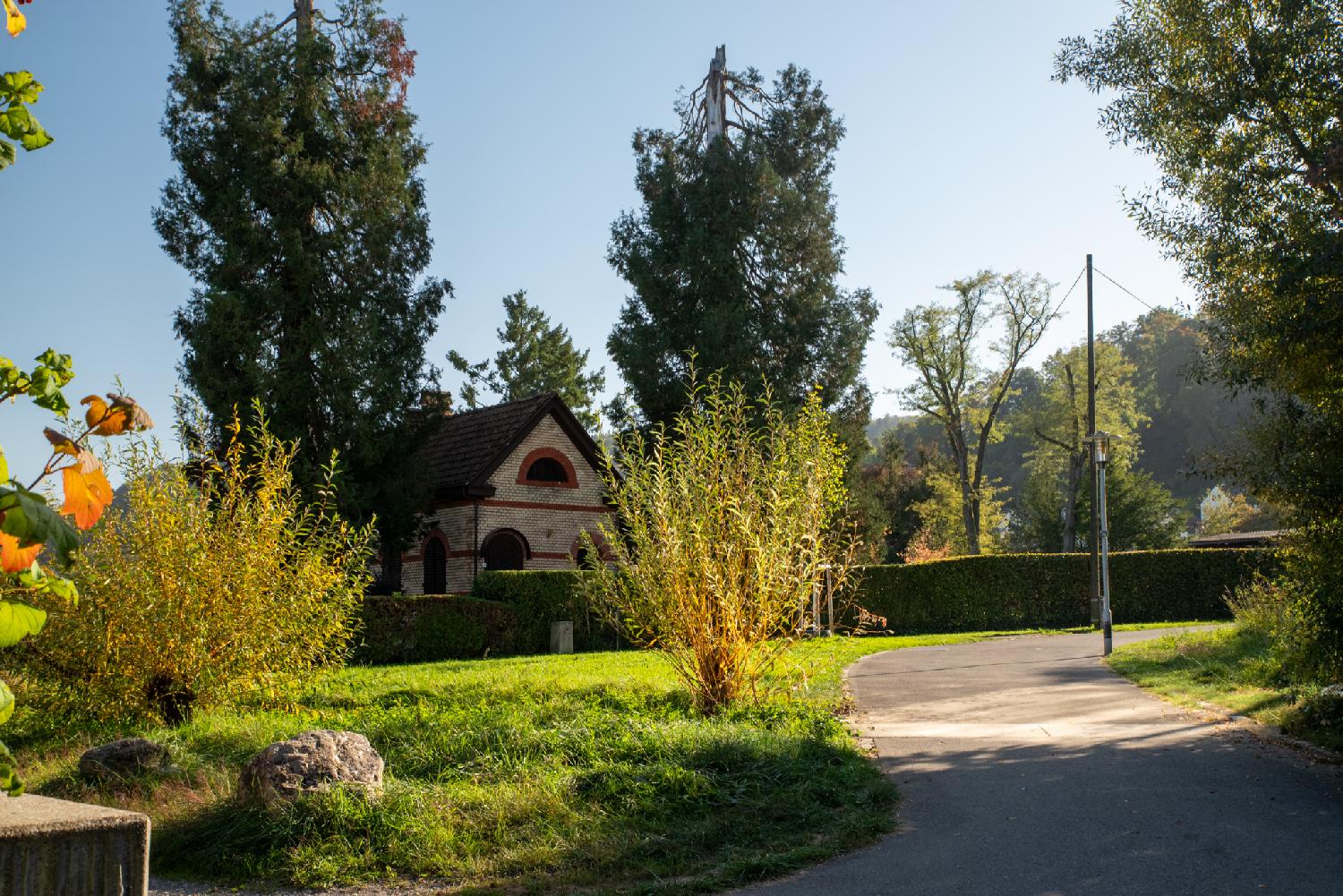 Algierstrasse in Zürich, where it starts to be a pedestrian path. A small brick building between two trees, more trees in the background, a narrow asphalted path.