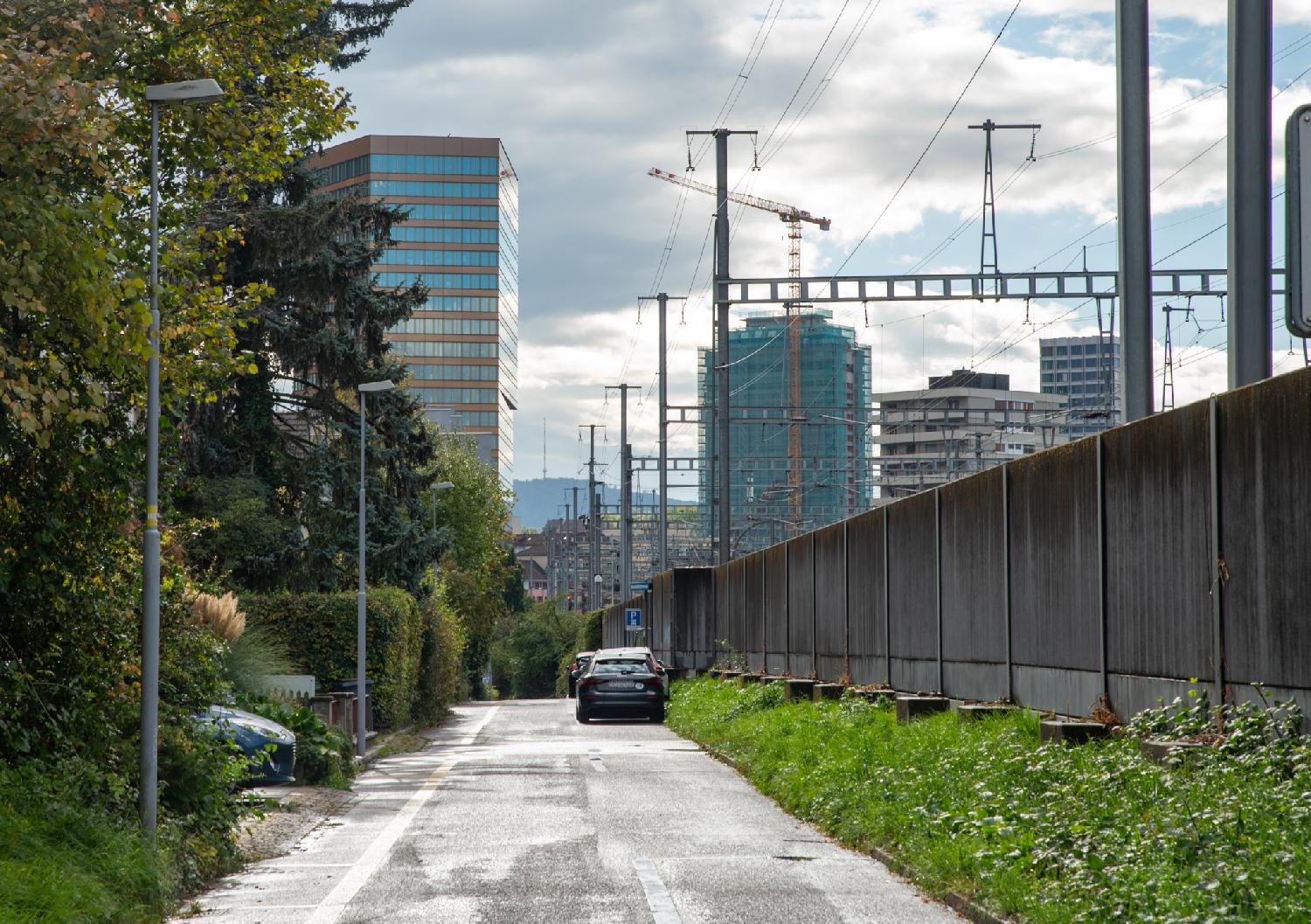 View over Oerlikon from Allmannstrasse: a narrow street with, on the right, a wooden fence separating the street from the train track and, in the background, high rises buildings and a far-away hill.