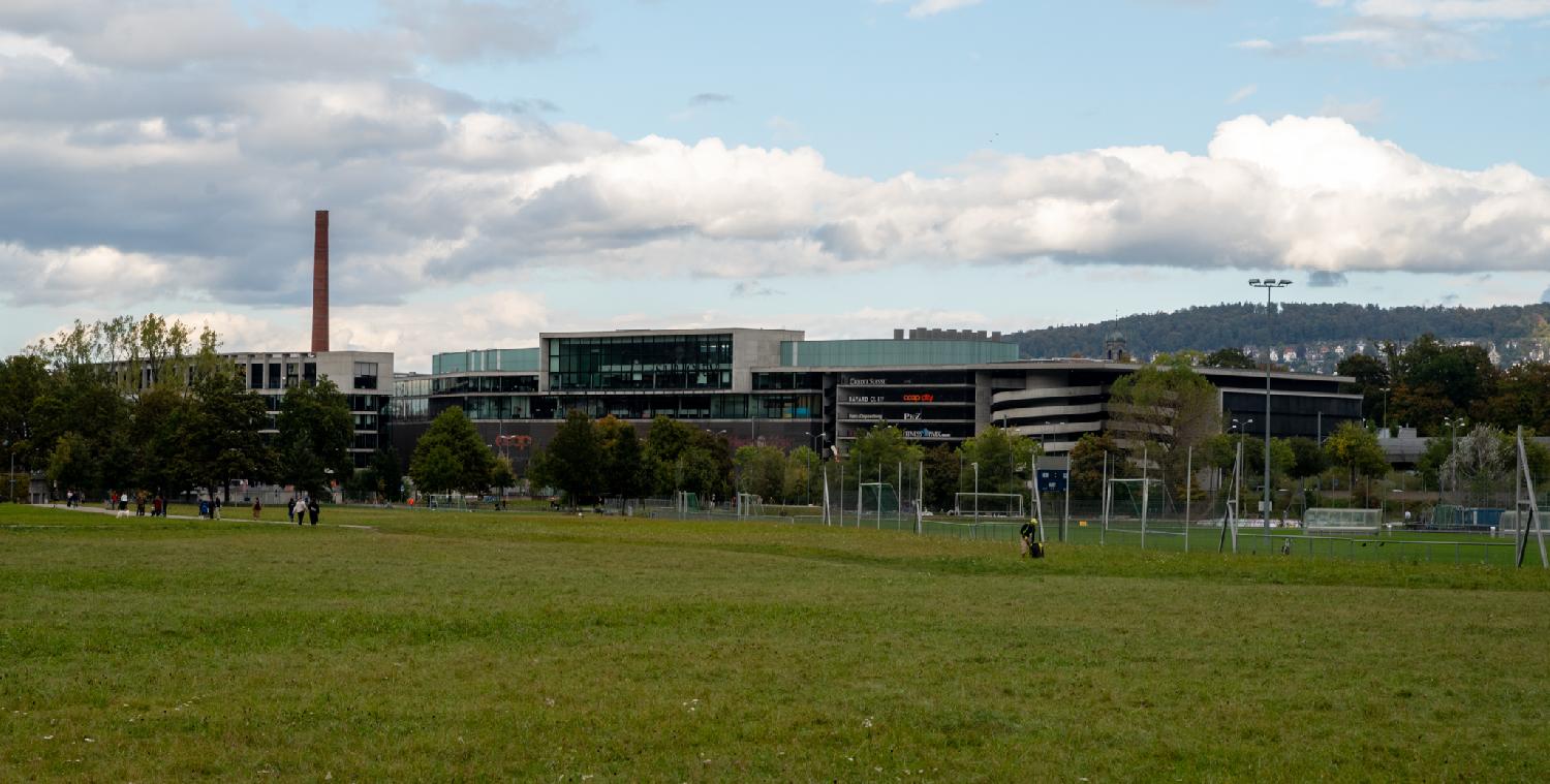 Sihlcity mall seen from Allmendweg in Zürich: a mall with buildings and a spiral parking lot, as well as a high brick chimney, with a grass meadow in the foreground