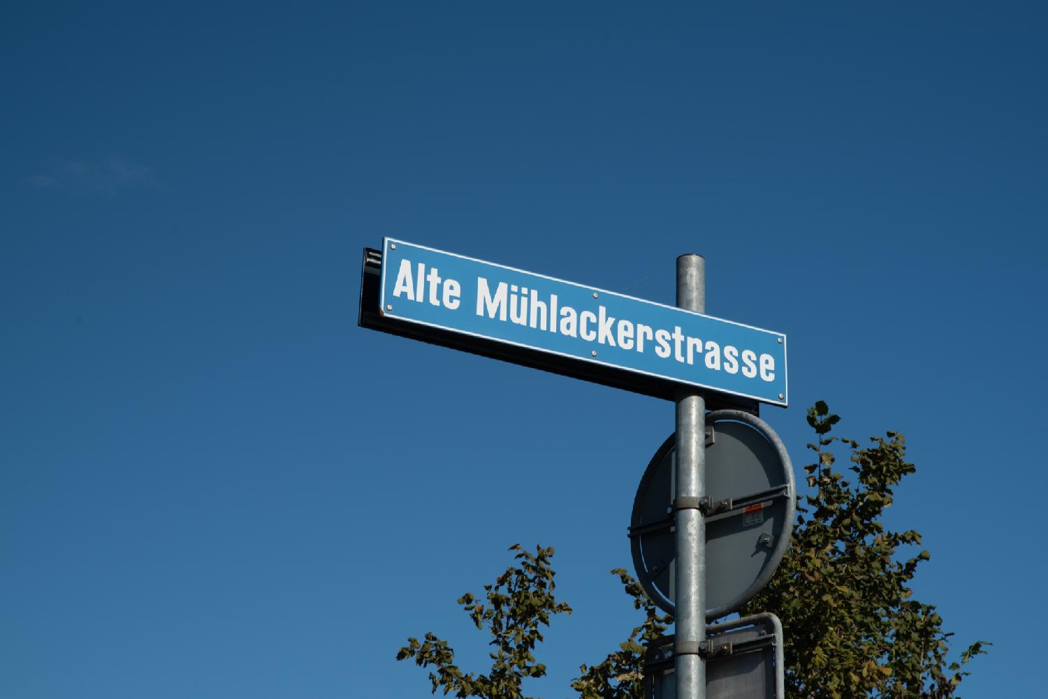 Street sign for Alte Mühlackerstrasse in Zürich: a blue sign on a greay pole, over a blue sky.