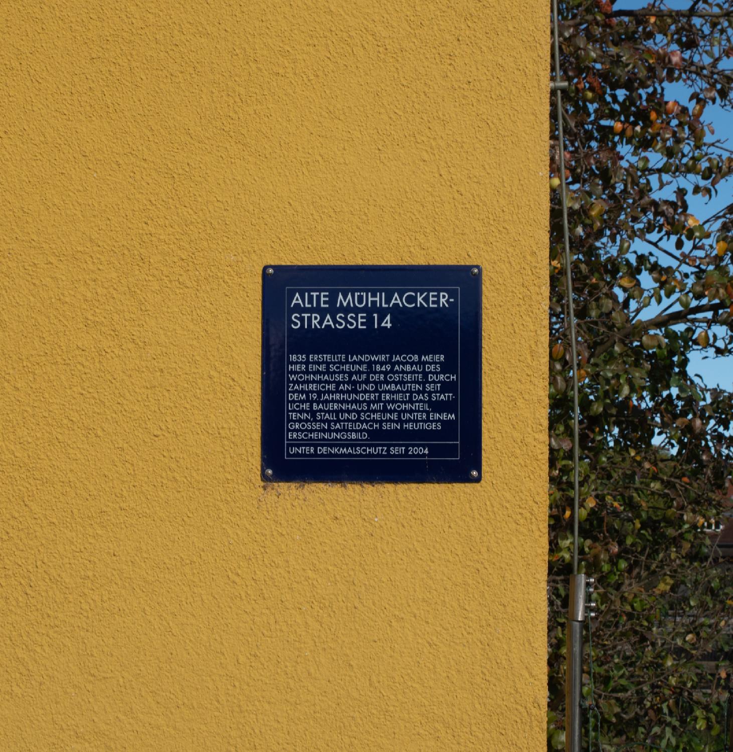 Denkmalschutz plate for Alte Mühlackerstrasse 14. A dark blue metallic plate on a yellow wall. The German text reads: "Alte Mühlackerstrasse 14 - 1835 erstellte Landwirt Jacob Meier hier ein e Scheune. 1849 Anbau des Wohnhauses auf der Ostseite. Durch Zahlreiche An- und Ambauten seit dem 19. Jahrhundert erhielt das stattliche Bauernhaus mit Wohnteil, Tenn, Stall und Scheune unter einem grossen Satteldach sein heutiges Ercheinungsbuild. Unter Denkmalschutz seit 2004"