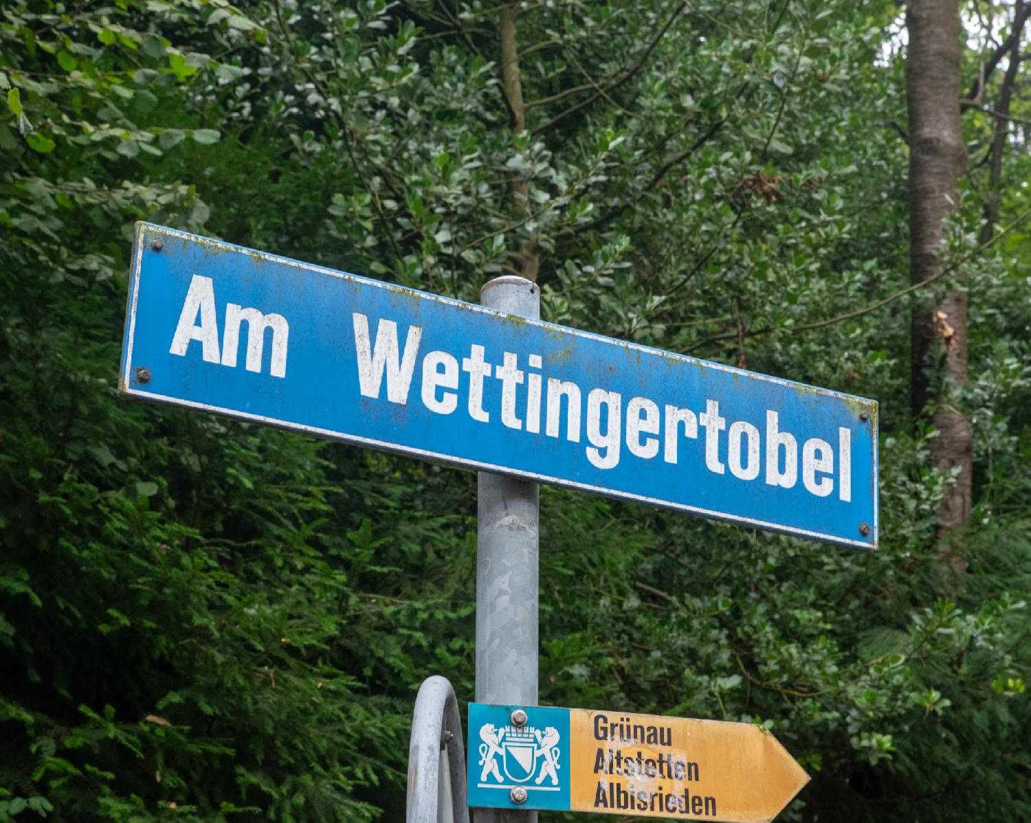 A blue "Am Wettingertobel" street sign above a yellow hiking sign to Grünau, Altstetten and Albisrieden, in front of dense trees.