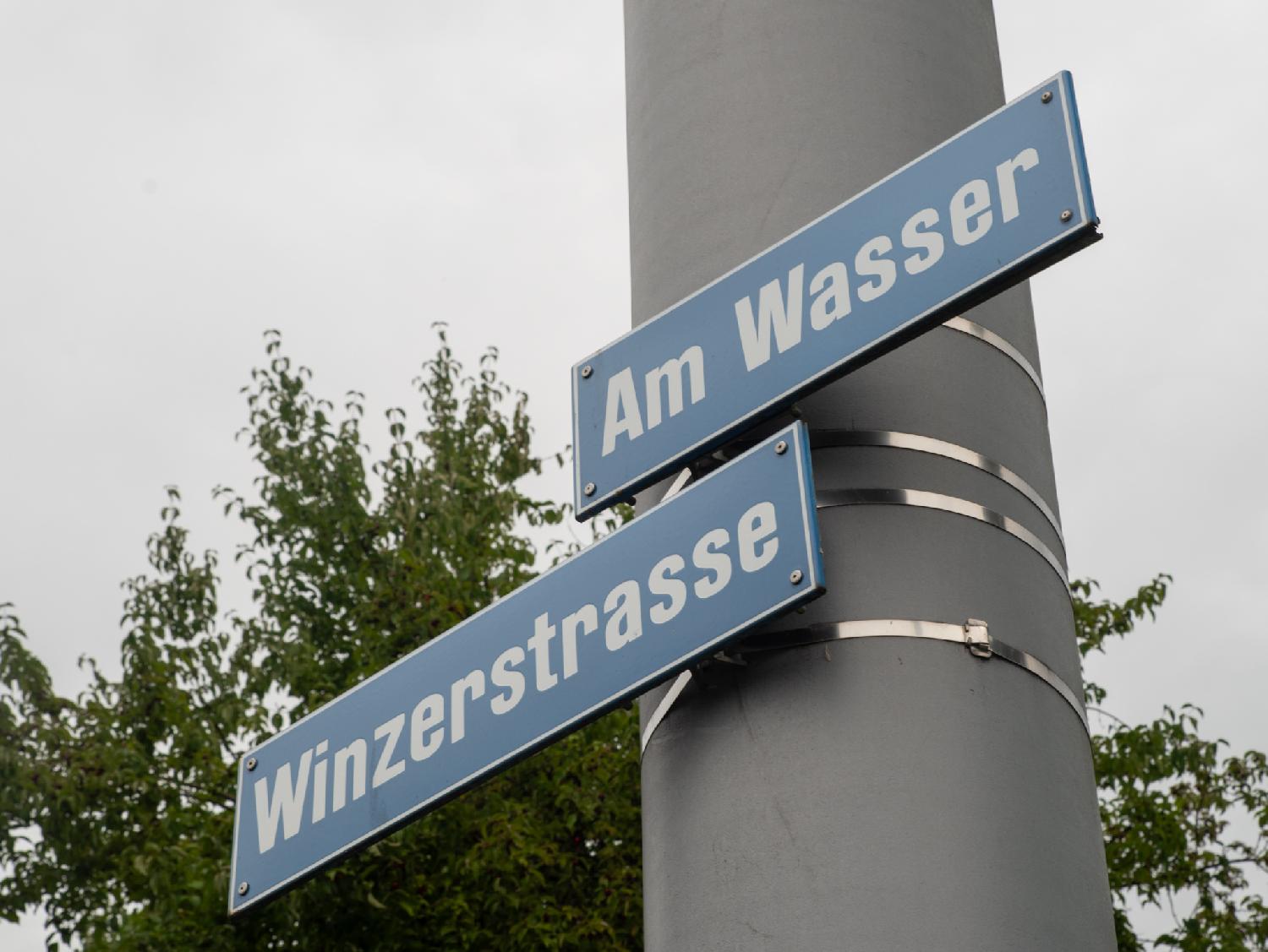 Blue "Winzerstrasse" and "Am Wasser" street signs attached to a large grey pole.