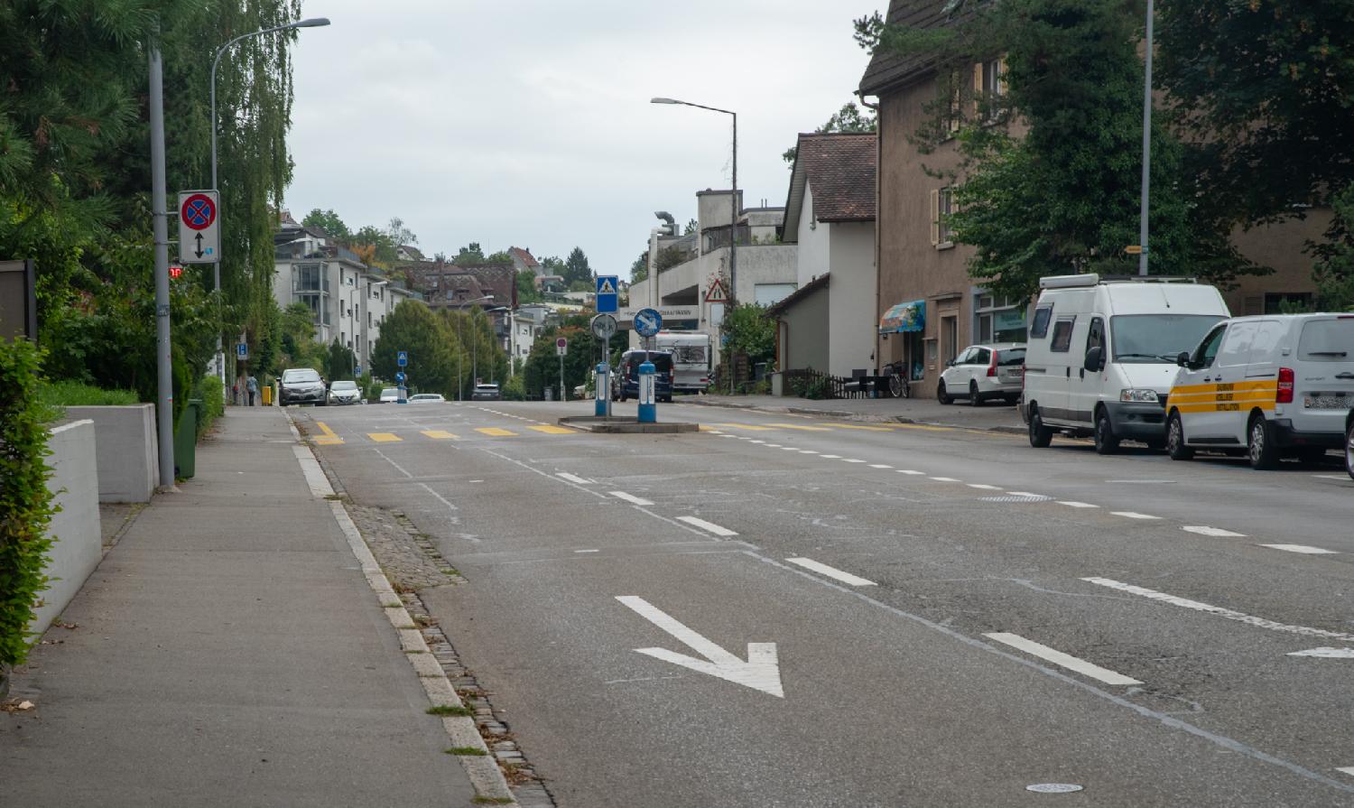 A 3-lane, 2-way street with a zebra crossing, cars parked on each side, and 2-3-story residential buildings along the street.