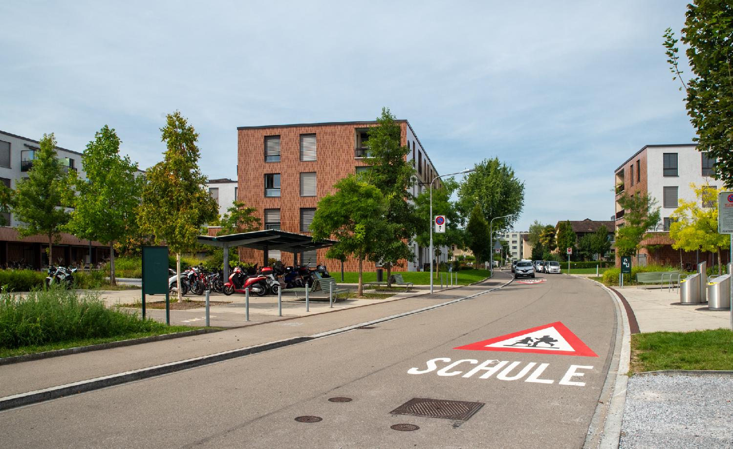 Am Glattbogen in Zürich: a two-way residential street with 4-story buildings, a "Schule" (School) sign painted on the road, a 2-wheel vehicle parking lot and some trees.