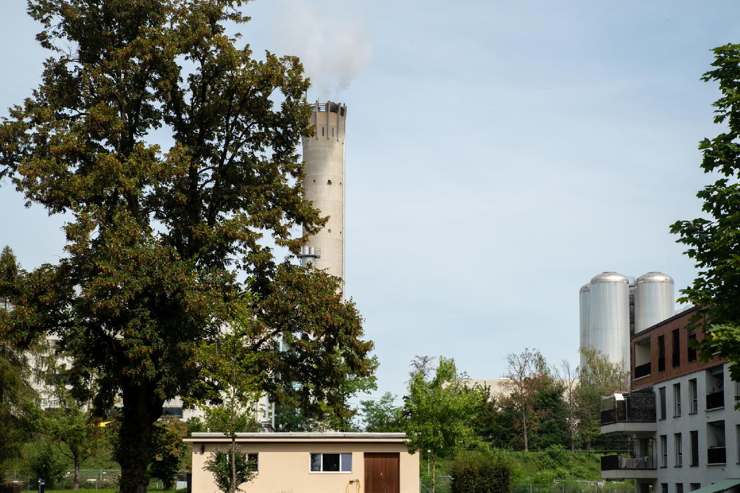 Hagenholz seen from Am Glattbogen: a concrete chimney and metallic silos, behind buildings and a large tree.