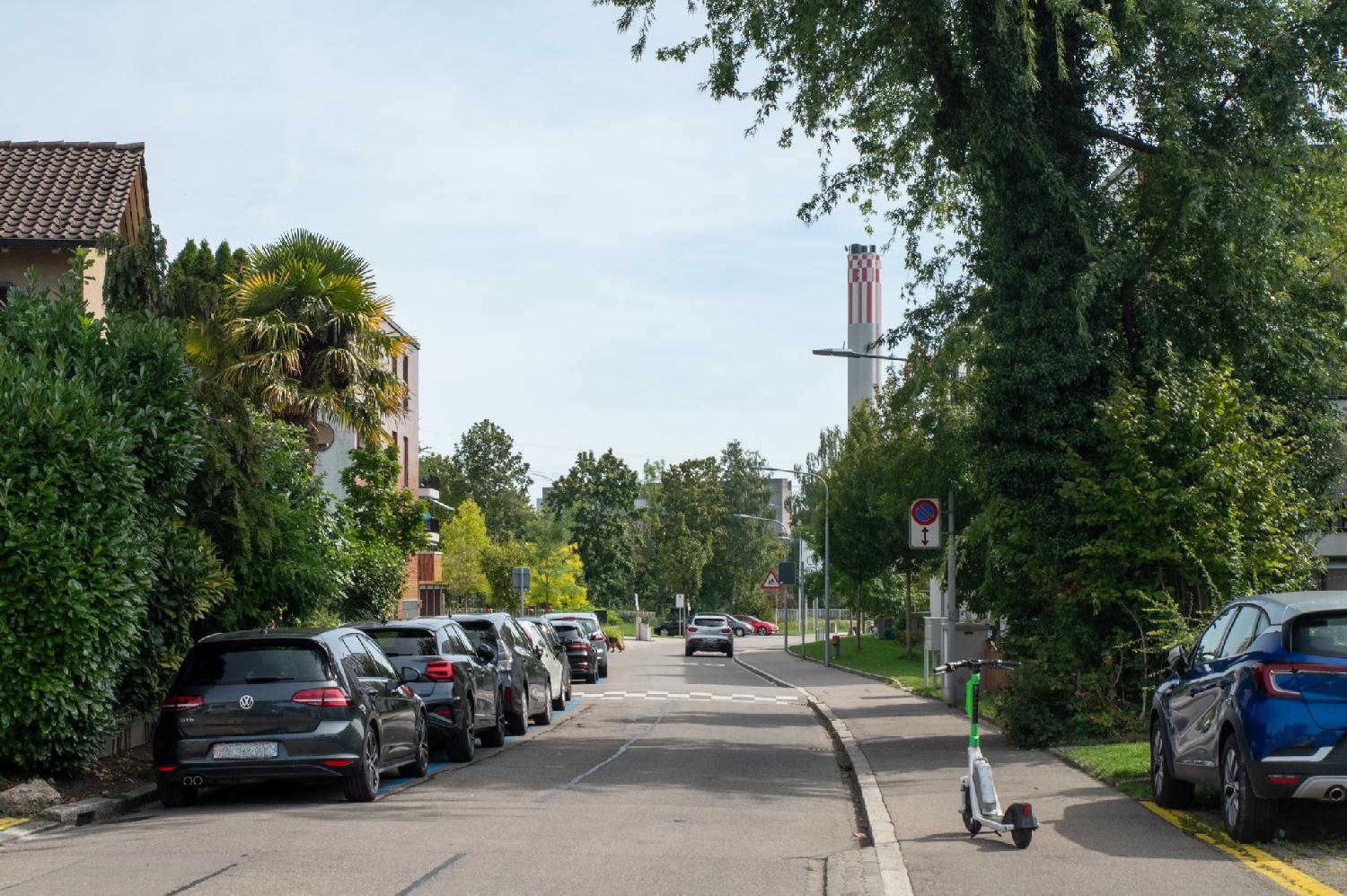 Heizkraftwerk Aubrugg seen from Am Glattbogen: a residential street with trees and 3-4 story buildings, and a chimney with red and white checker pattern on top in the background.