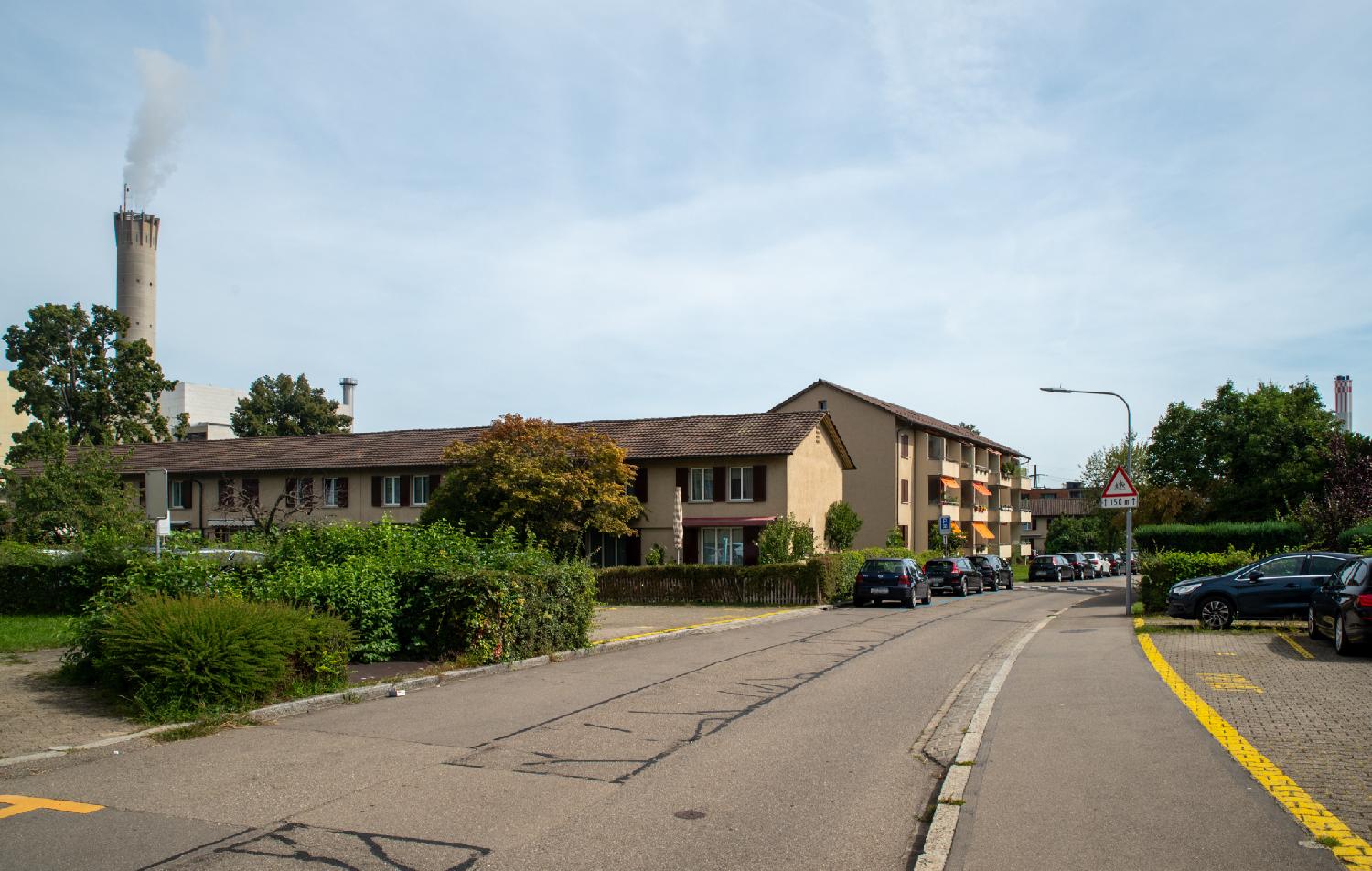 A residential street with 2-3 story buildings and cars parked on each side, with two industrial chimneys visible on each side of the background