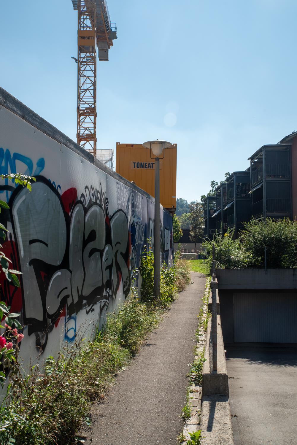 A very narrow path between a construction fence (with a lot of graffiti) on the left and 3-story buildings on the right behind a descending garage entrance.