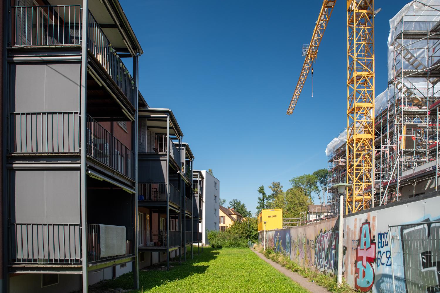 A very narrow path between 3-story black buildings on the left and a construction site with fences and a yellow crane on the right.