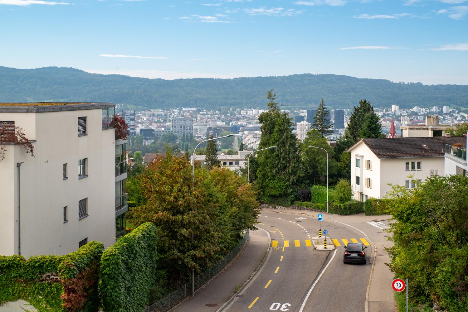 A two-way-street seen from the bridge above it, with the city of Zürich and the Uetliberg (hilly forest) in the background.