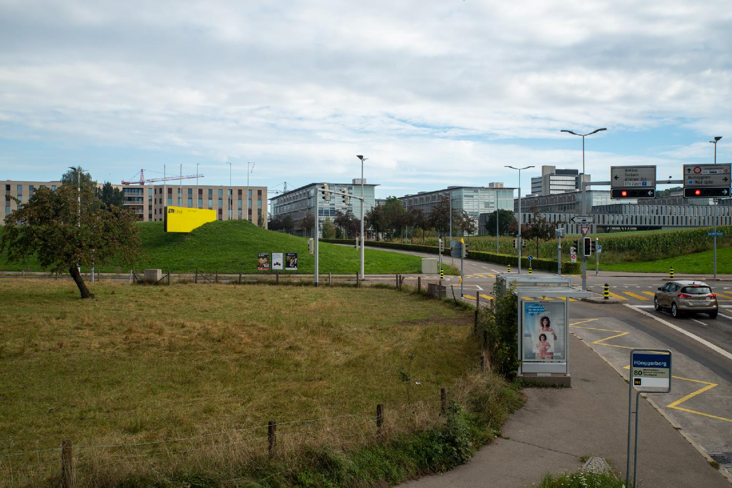 ETH Hönggerberg campus, seen from Hönggerbergstrasse: 4-5 stery buildings with varying amount of glass, behind a street and a small meadow. There's a large bright yellow ETHZürich sign in front of the buildings, and a Hönggerberg bus stop in the foreground.