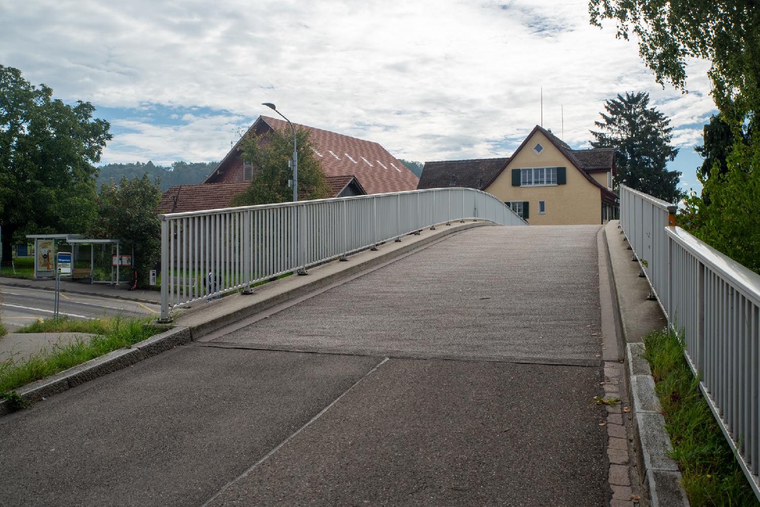 Zürich Hönggerbergstrasse bridge: a curved pedestrian bridge with metalic fences on top of a road and a bus stop, with a couple of buildings in the background.