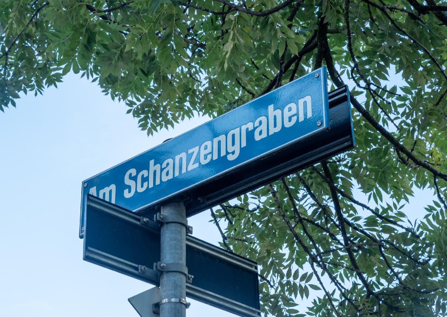 Blue "Am Schanzengraben" street sign, with another sign (seen from behind) perpendicular to it, and tree branches and leaves in the background.