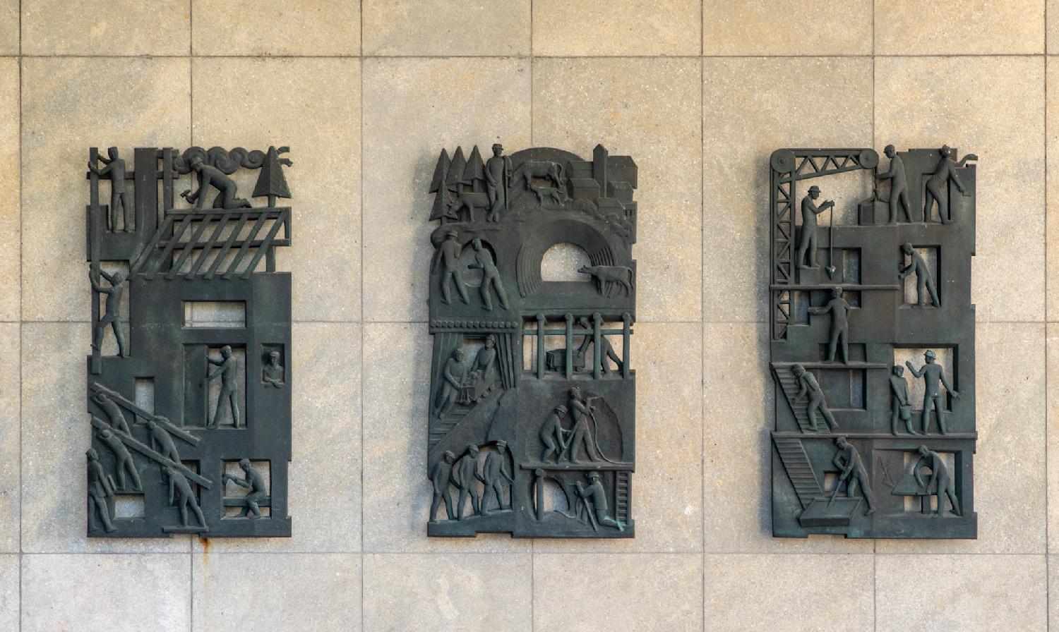Metallic reliefs depicting industrial scenes: men moving beams, using a crane, moving wagons in tunnels. The reliefs are hollow and fixed on a beige stone wall.